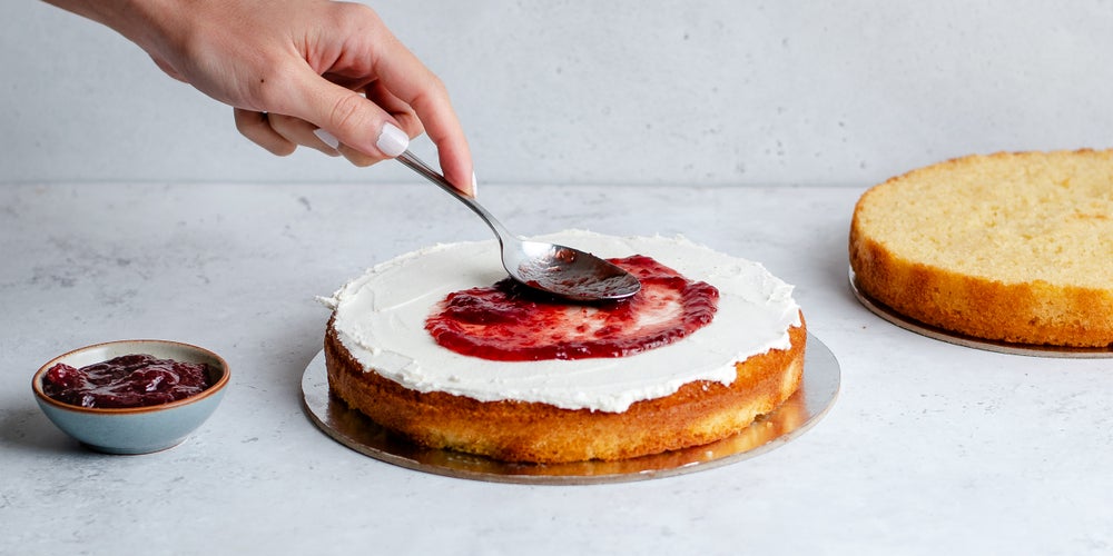 Sponge cake cover in buttercream with a hand spooning jam on top.
