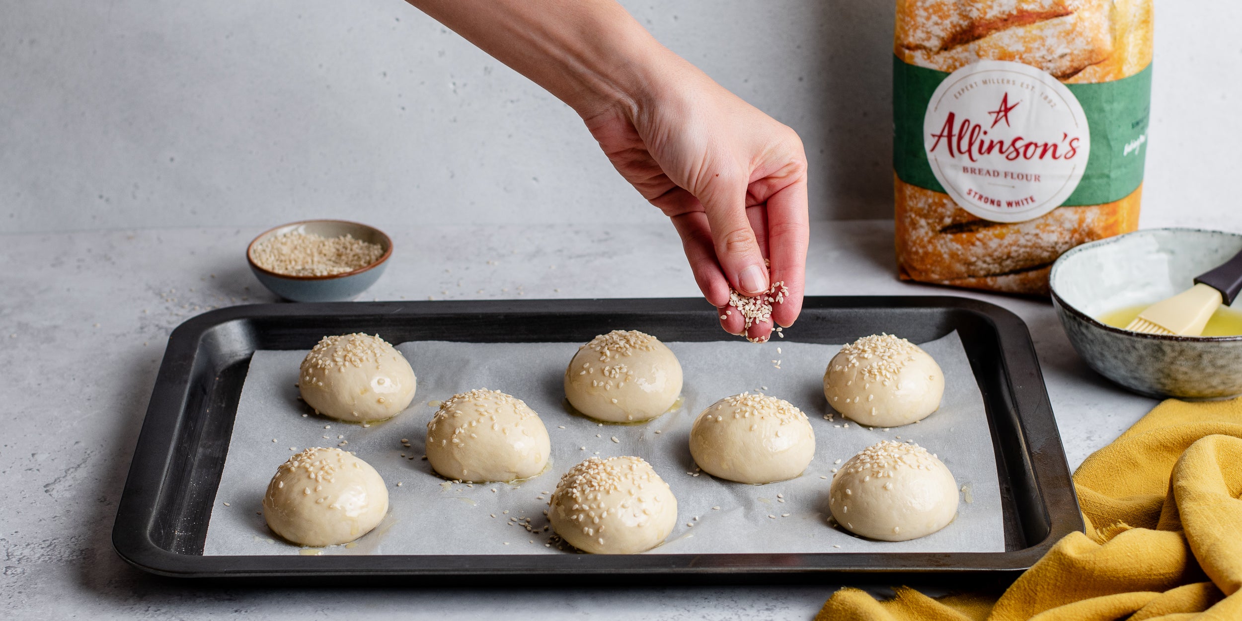 Hand sprinkling sesame seeds on top of bread buns on baking tray with flour pack in shot