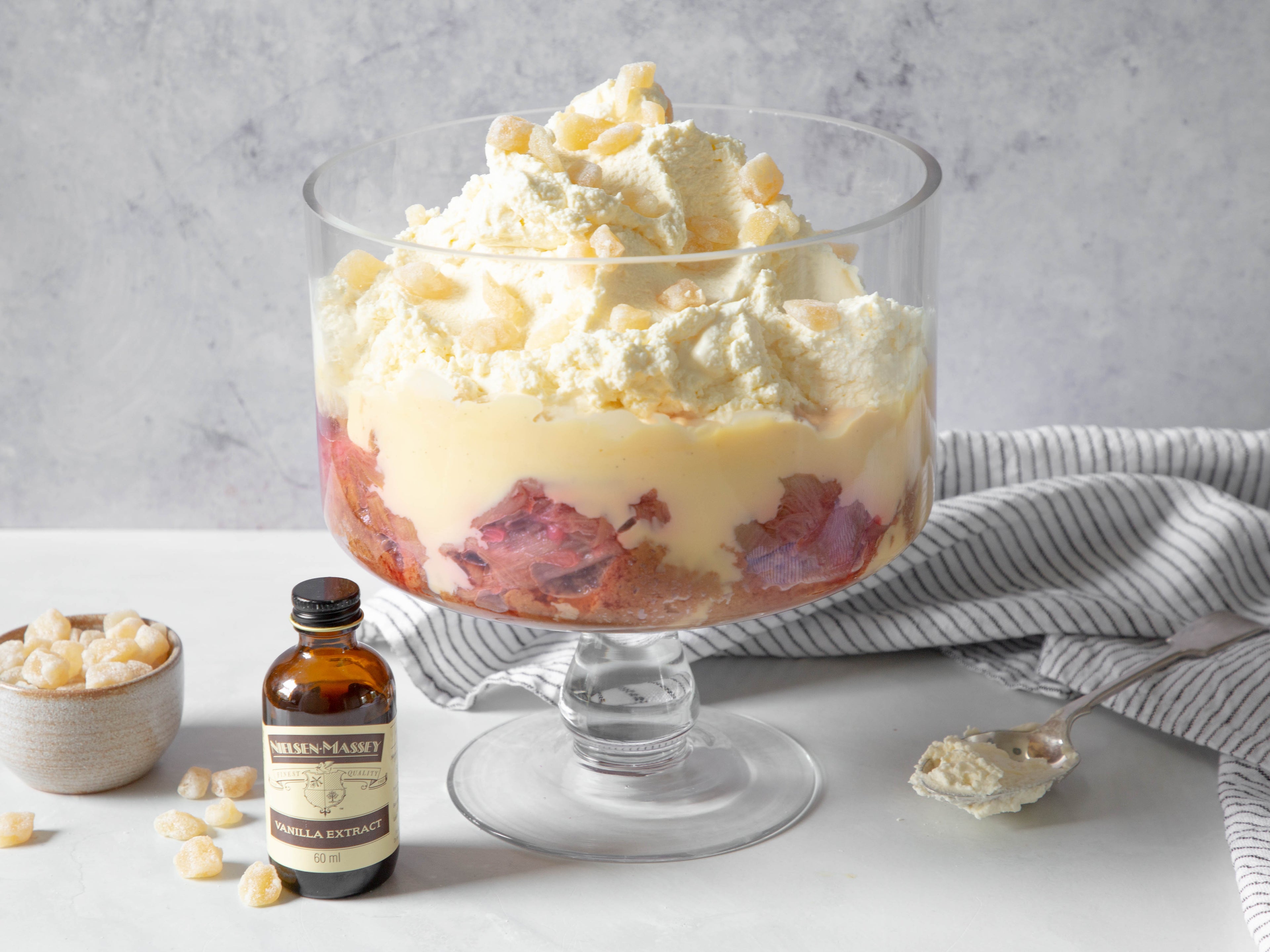 Glass trifle bowl filled with trifle, a striped teatowel in the background. A spoon covered in cream, a vanilla bottle and small bowl of nuts in the forefront