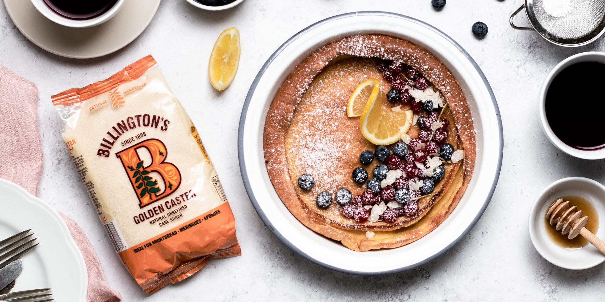 Stack of Dutch Baby Pancakes drizzled in honey, berries and lemon, next to a pack of Billington's Golden Caster Sugar 