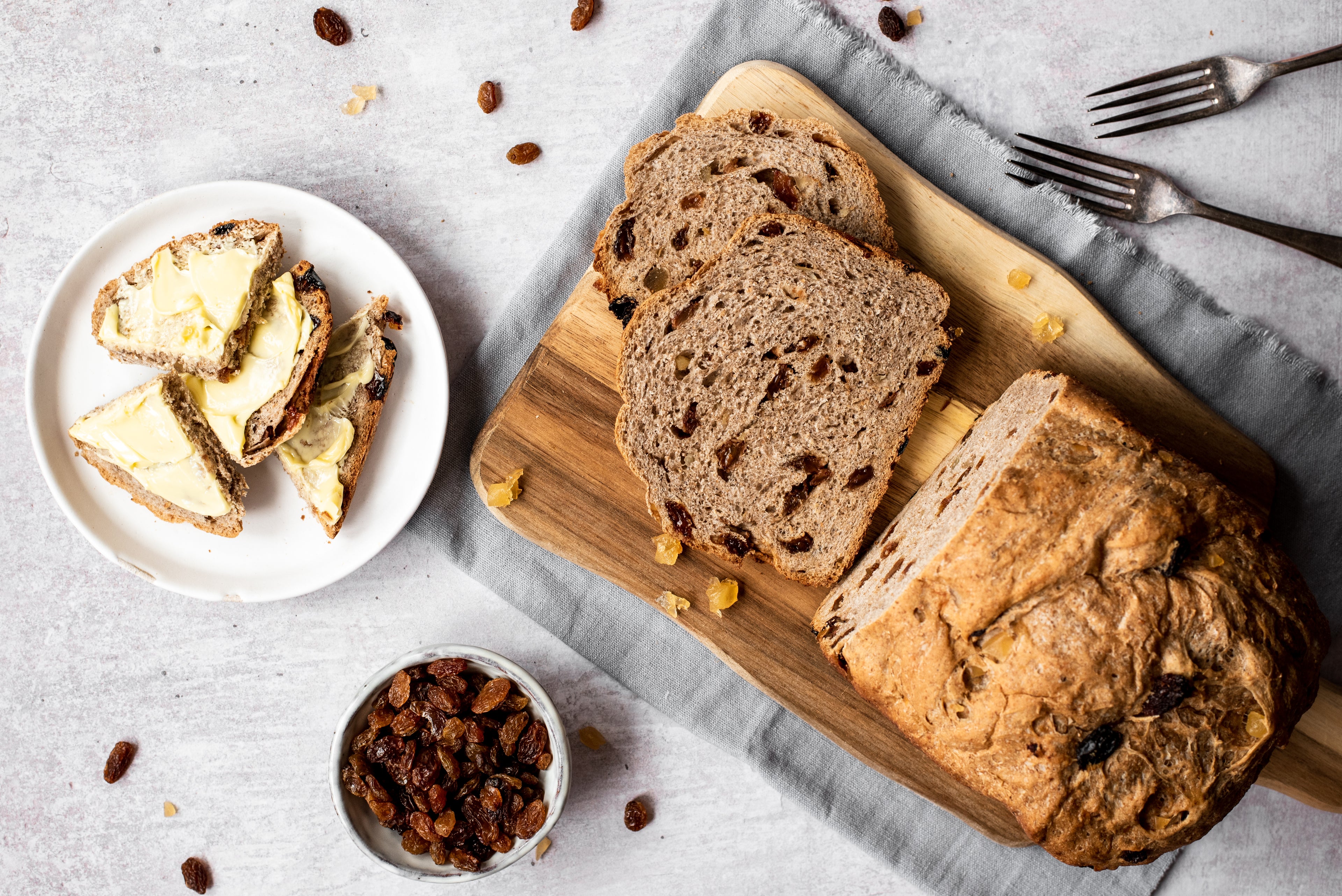 Overhead of fruit bread with slices cut. Plate with buttered slices. Bowl of sultanas. Two forks