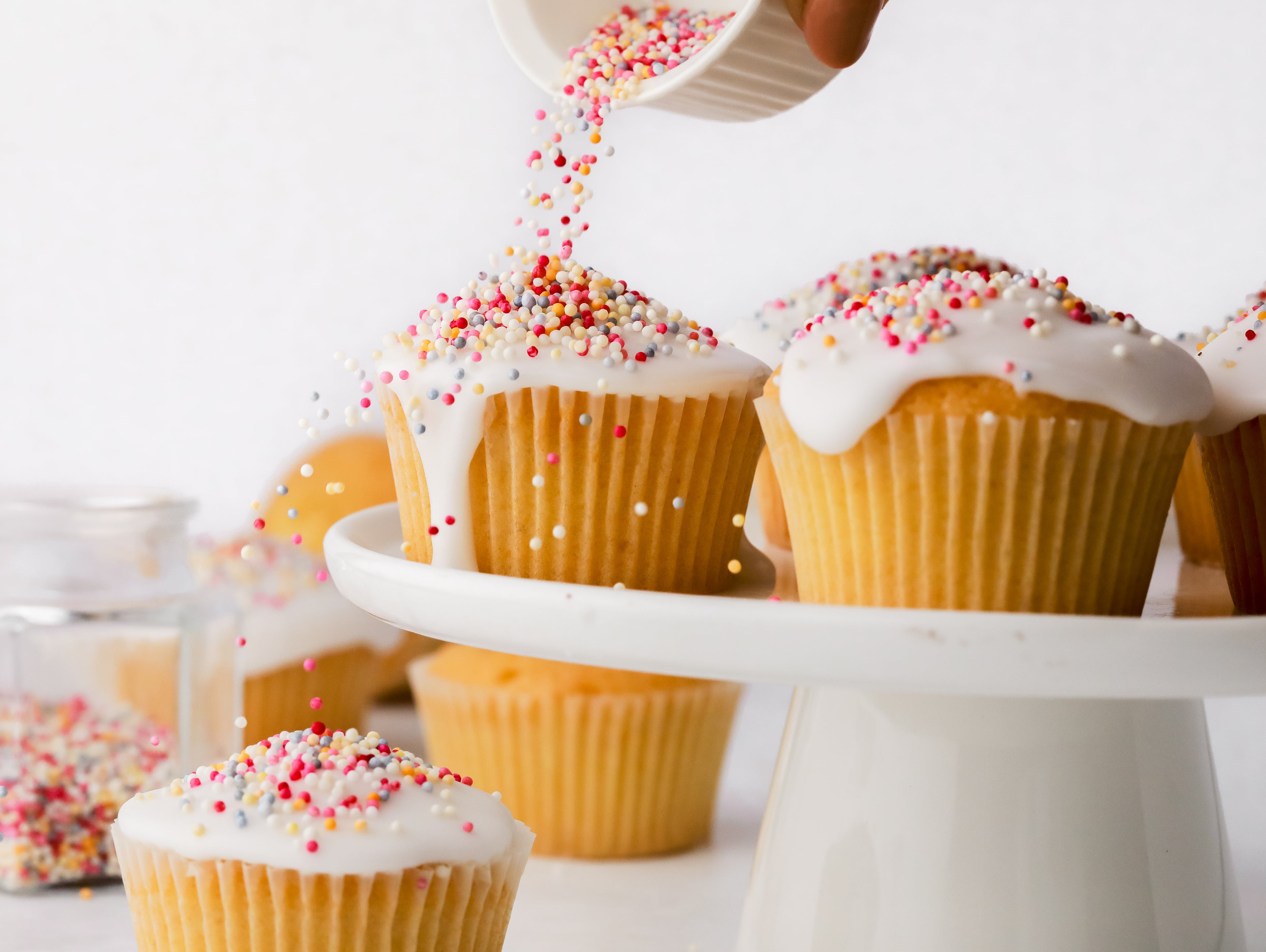 Sprinkles being poured on top of a fairy cake covered in white icing 