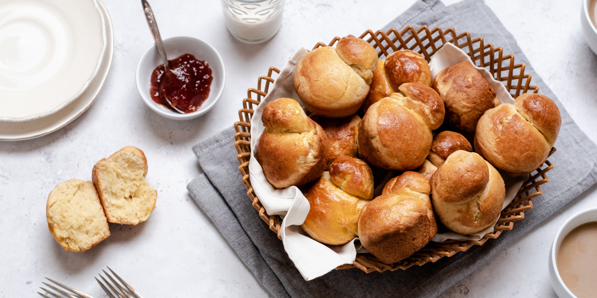 Batch of Brioche in a wicker basket, with a brioche torn in half showing the fluffy insides. Next to a bowl of jam and a glass of milk