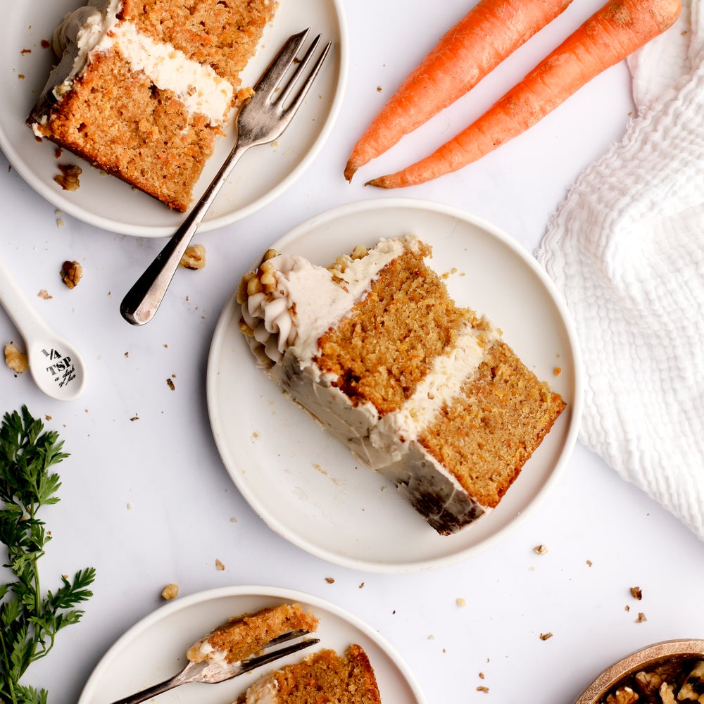 A slice of carrot cake topped with buttercream laid flat on a cream saucer and shot from above with some carrots to the side of the photo as if a messy kitchen scene.