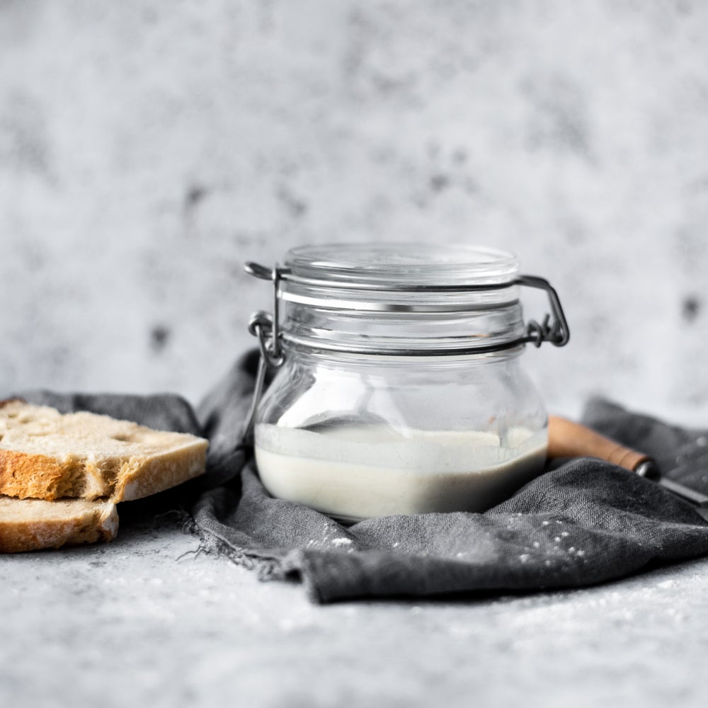 A glass jar of sourdough starter that resembles milk on a charcoal grey piece of fabric and next to some slices of bread.