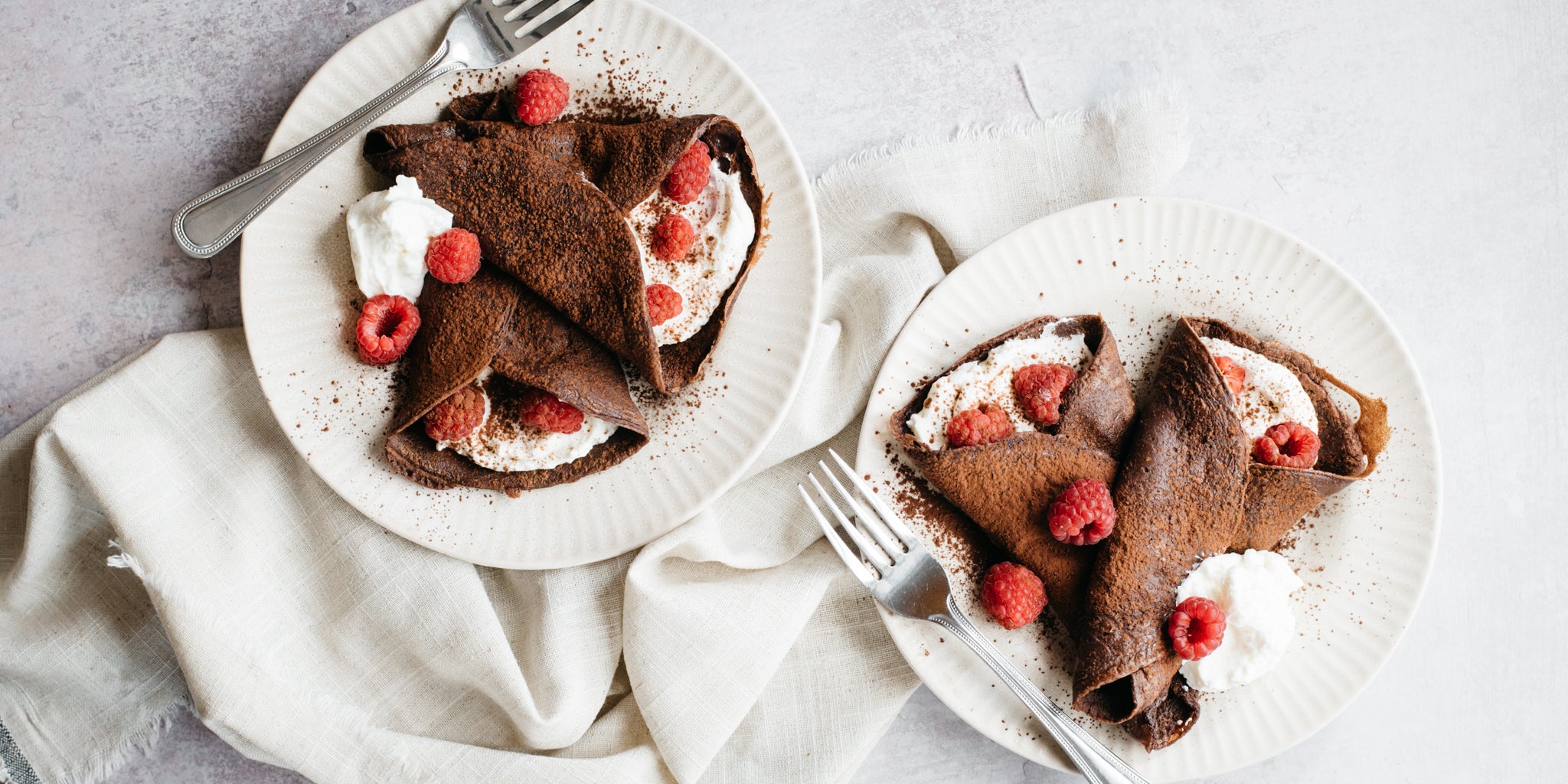 Two white plates with chocolate crepes, cream and raspberries