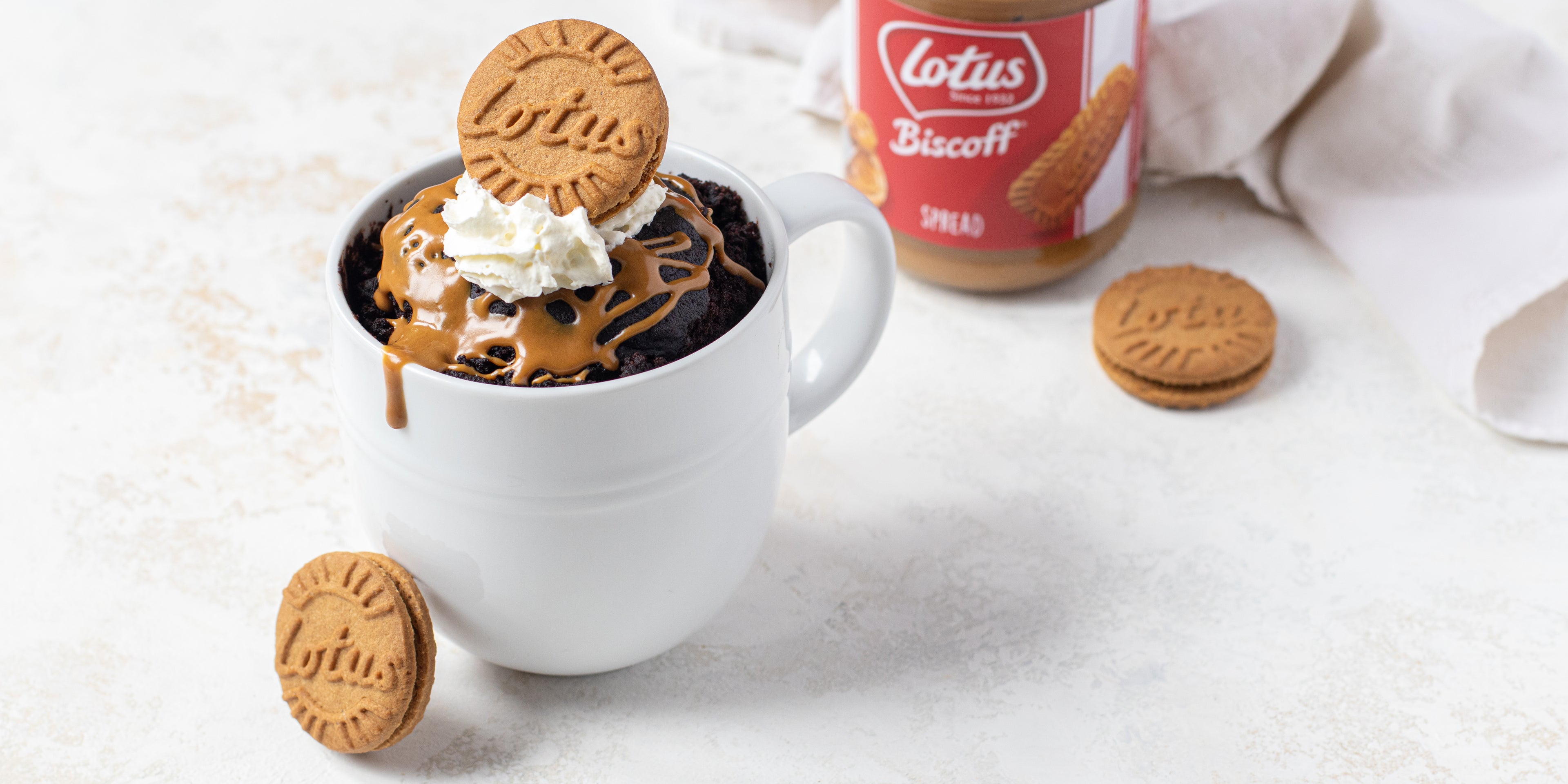 Close up of Chocolate Mug Cake customised with Lotus biscuits, drizzled in caramel sauce and a dollop of cream, with a jar of biscoff spread in the background