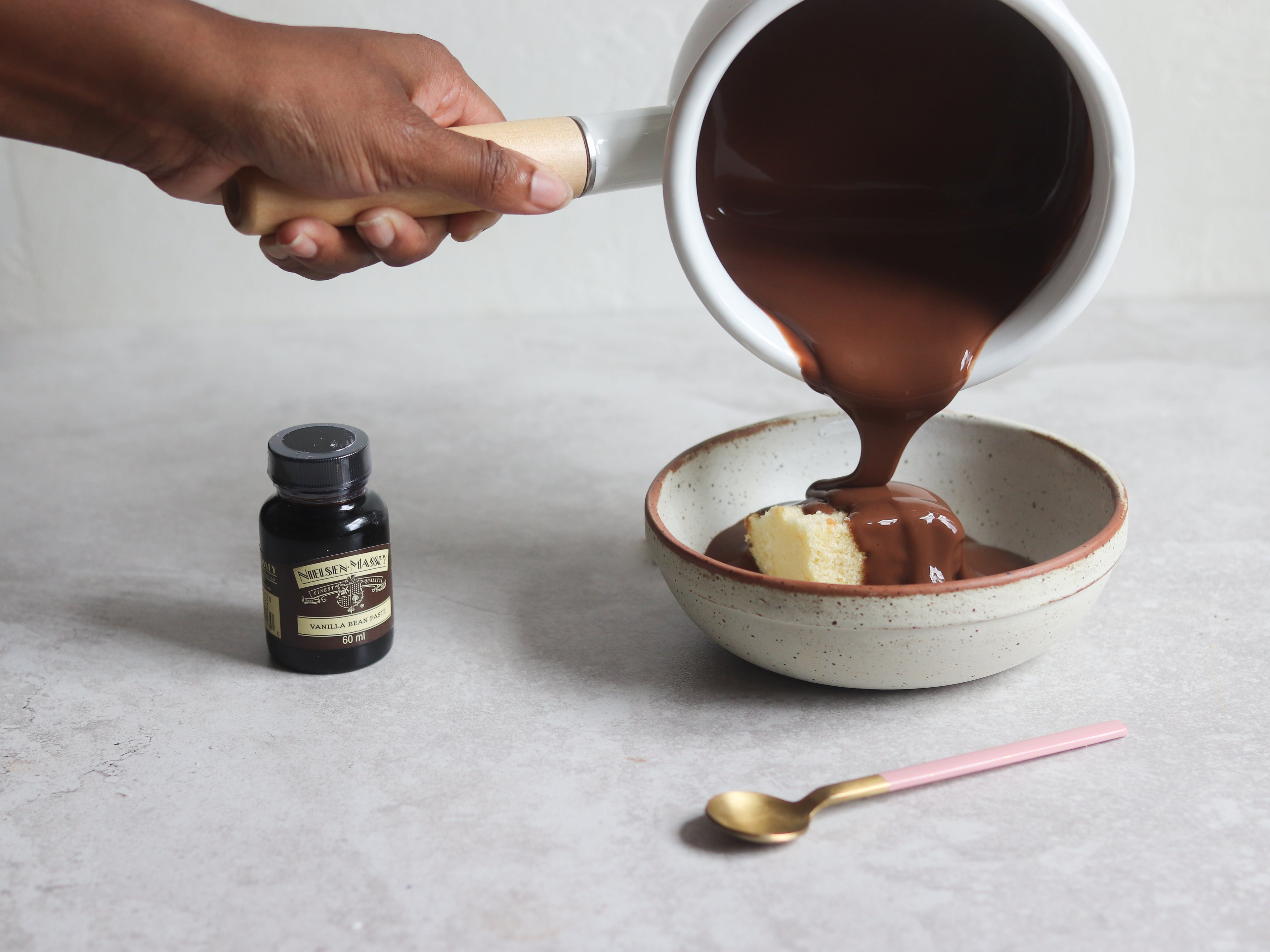 Chocolate Custard being poured over a sponge cake from a saucepan with a hand. Next to a bottle of Neilsen-Massey Vanilla Extract