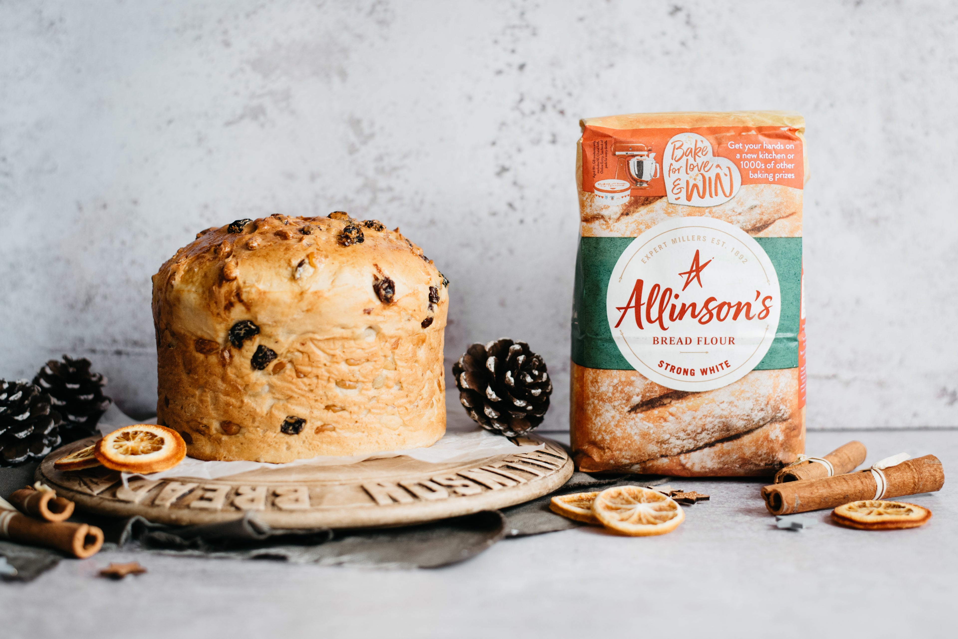 Golden Panettone sat on a wooden serving board, surrounded by cinnamon sticks, dried orange and pine cones, next to a bag of Allinson's Strong White Flour