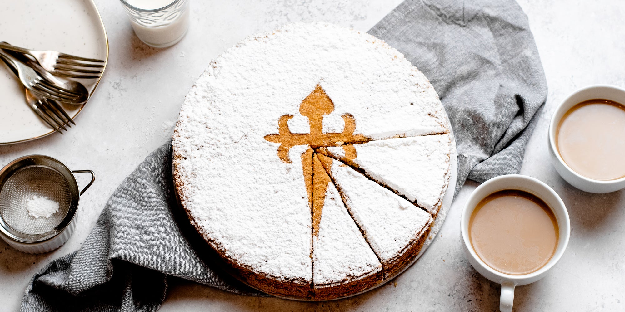 Tarta de Santiago with three slices cut out of it, served on a linen cloth, next to two cups of tea and cutlery ready to serve
