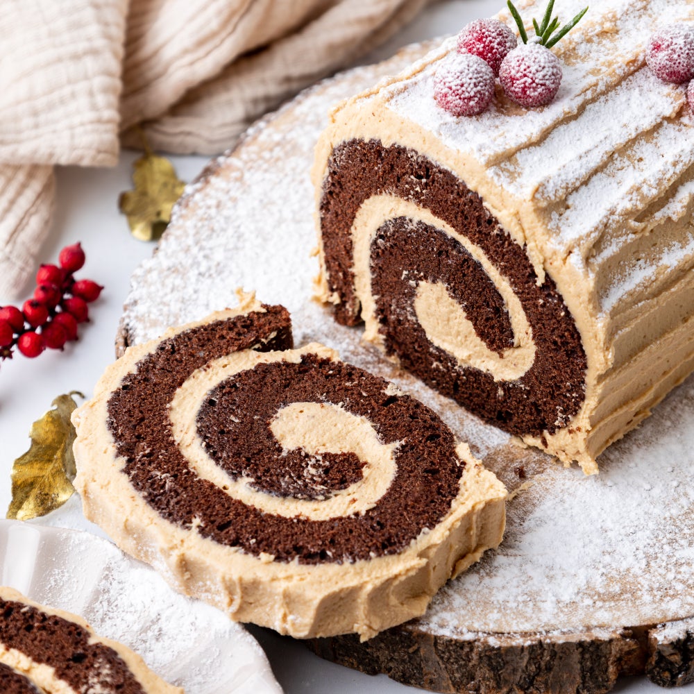 Biscoff Yule Log Cake with slice cut, decorated with festive accessories on a wooden plinth.