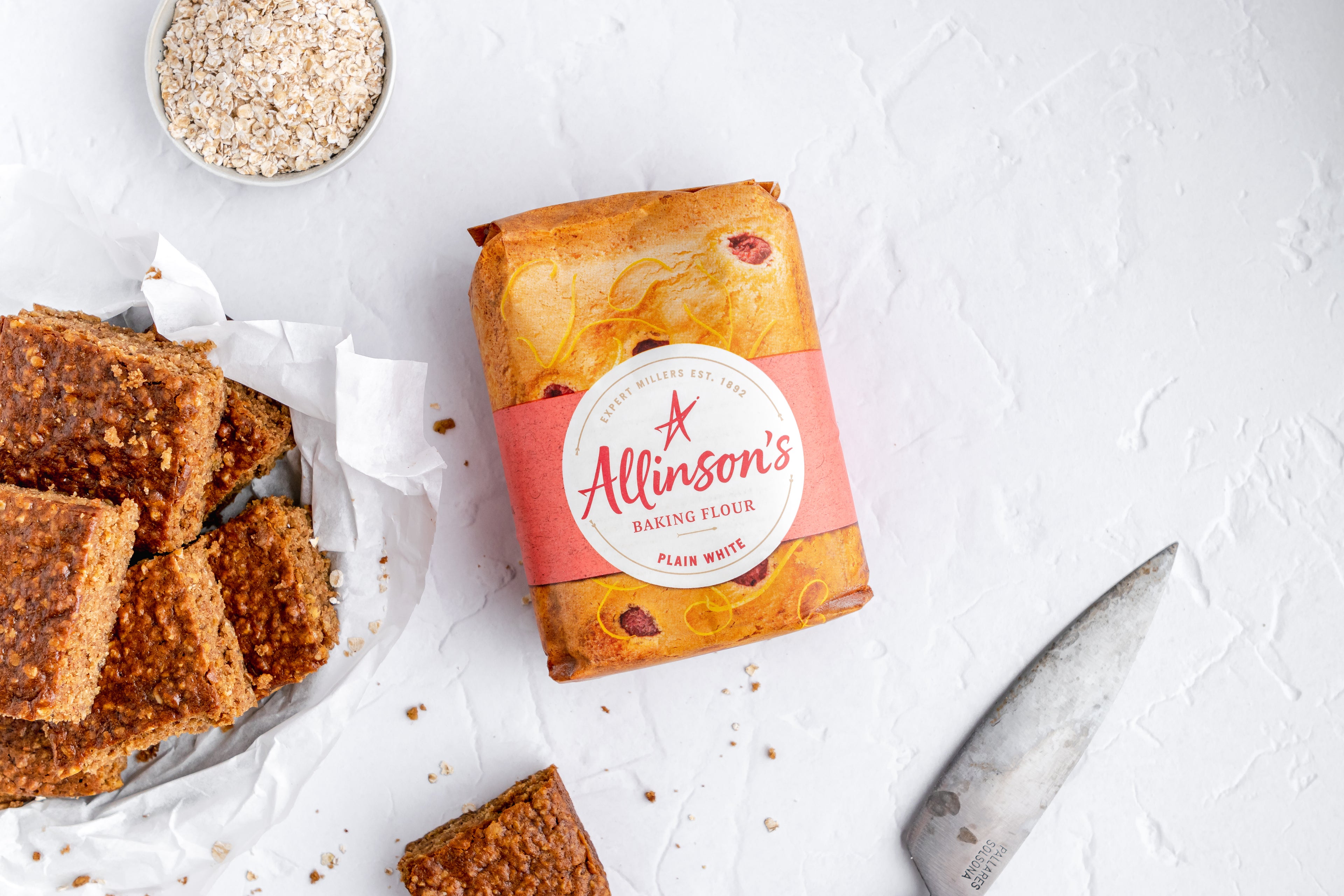 Sticky Yorkshire Parkin squares next to a small bowl of oats and Allinson's Plain White Flour