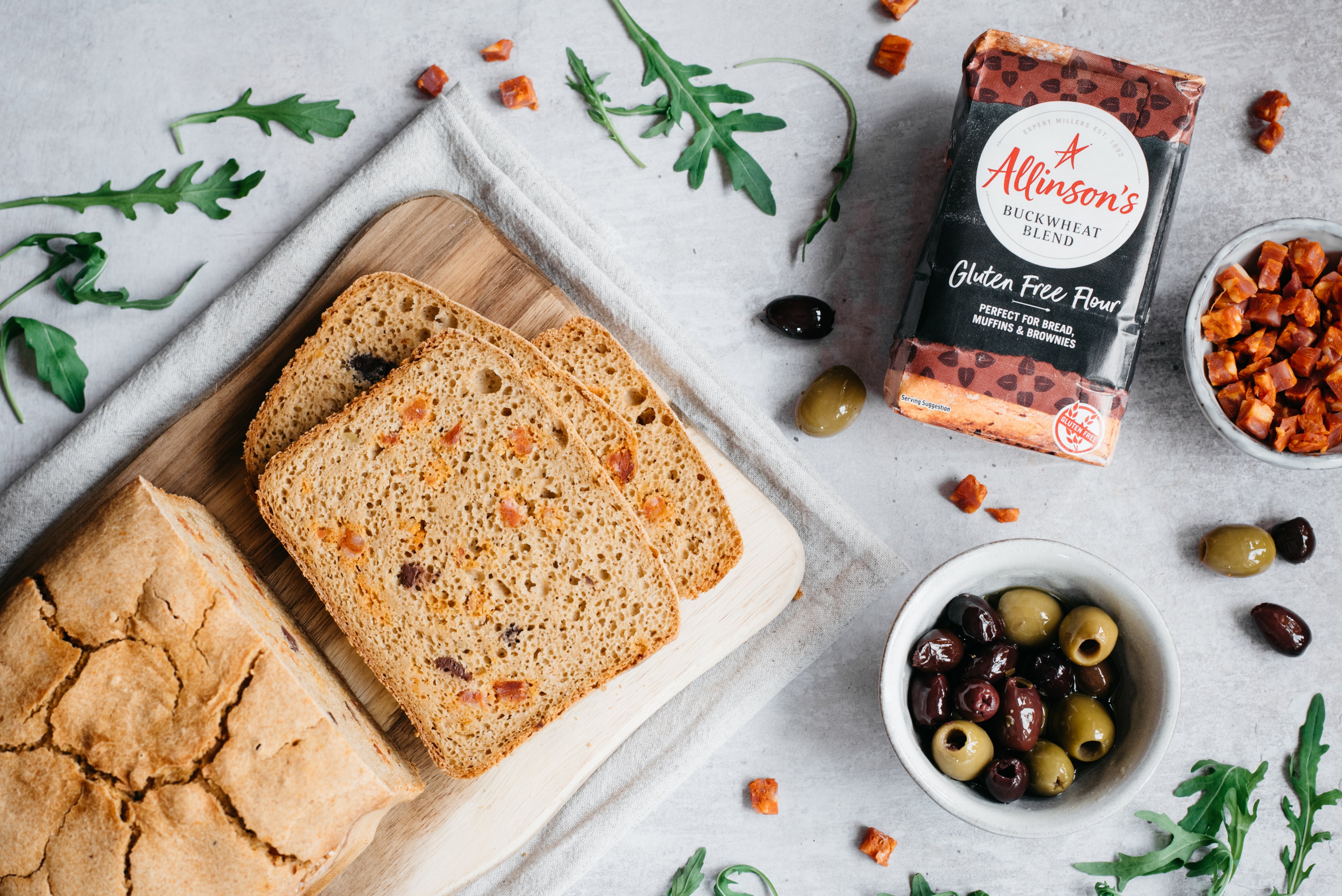 Over head shot of loaf of bread with slices cut, flour pack and two bowls of chorizo and olives