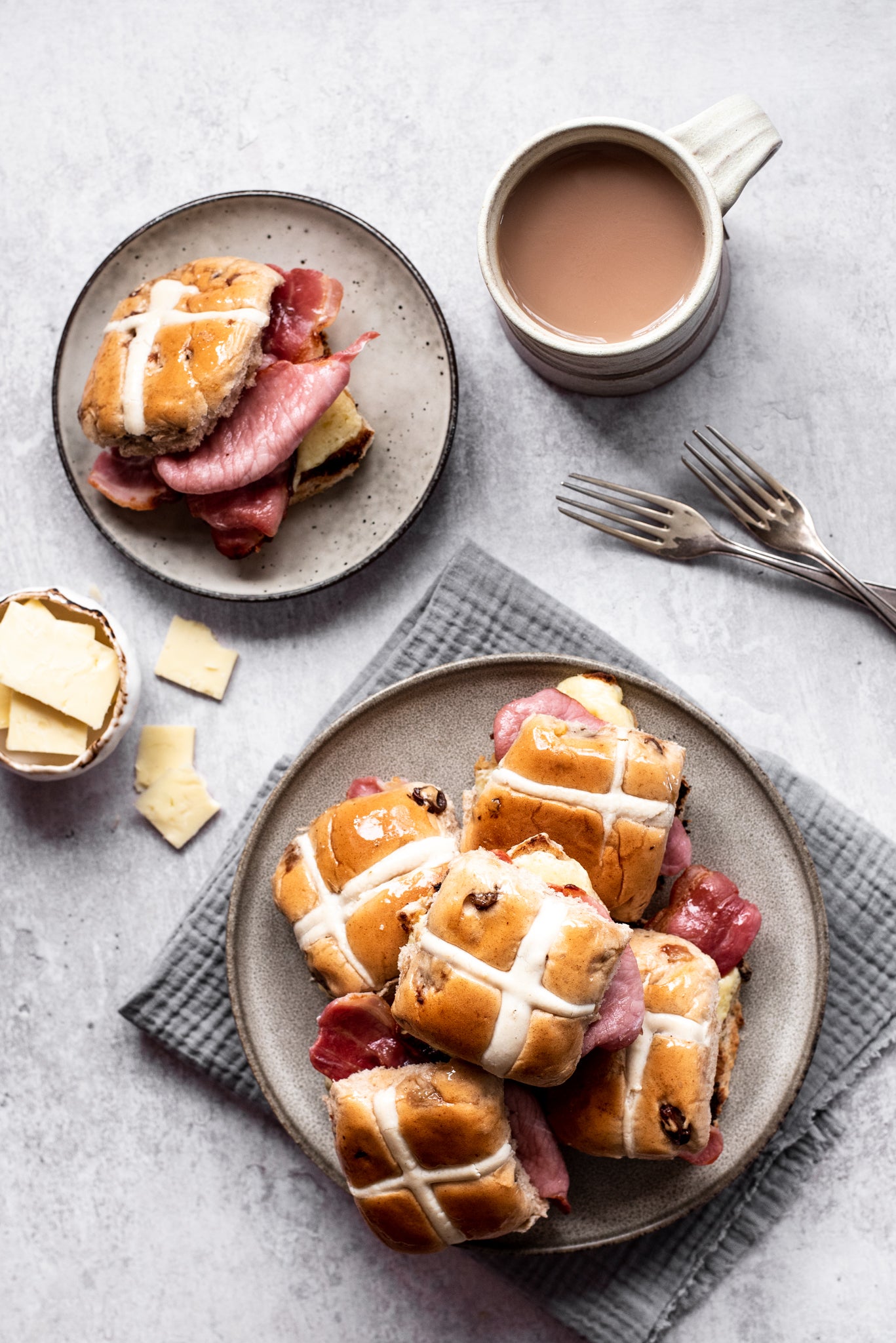 Plate of hot cross buns stacked up. Cup of tea. Two forks. Plate with hot cross bun and bacon