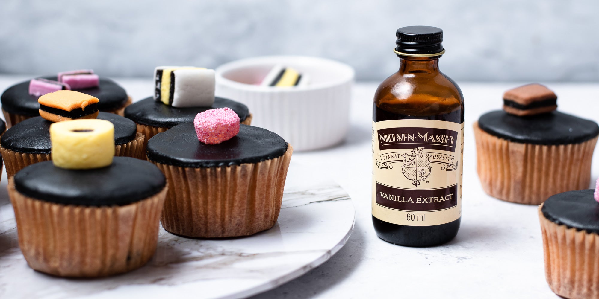 Cupcakes in brown cases, black fondant, topped with liquorice allsorts. Bowl of sweets in background. Vanilla bottle