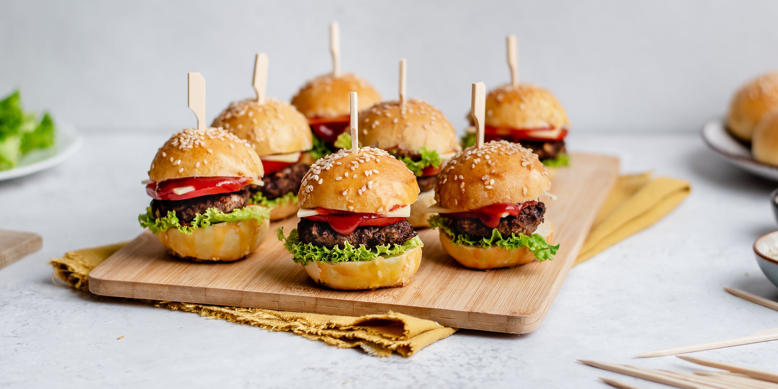 7 burgers lined up on wooden chopping board with skewers in the centre