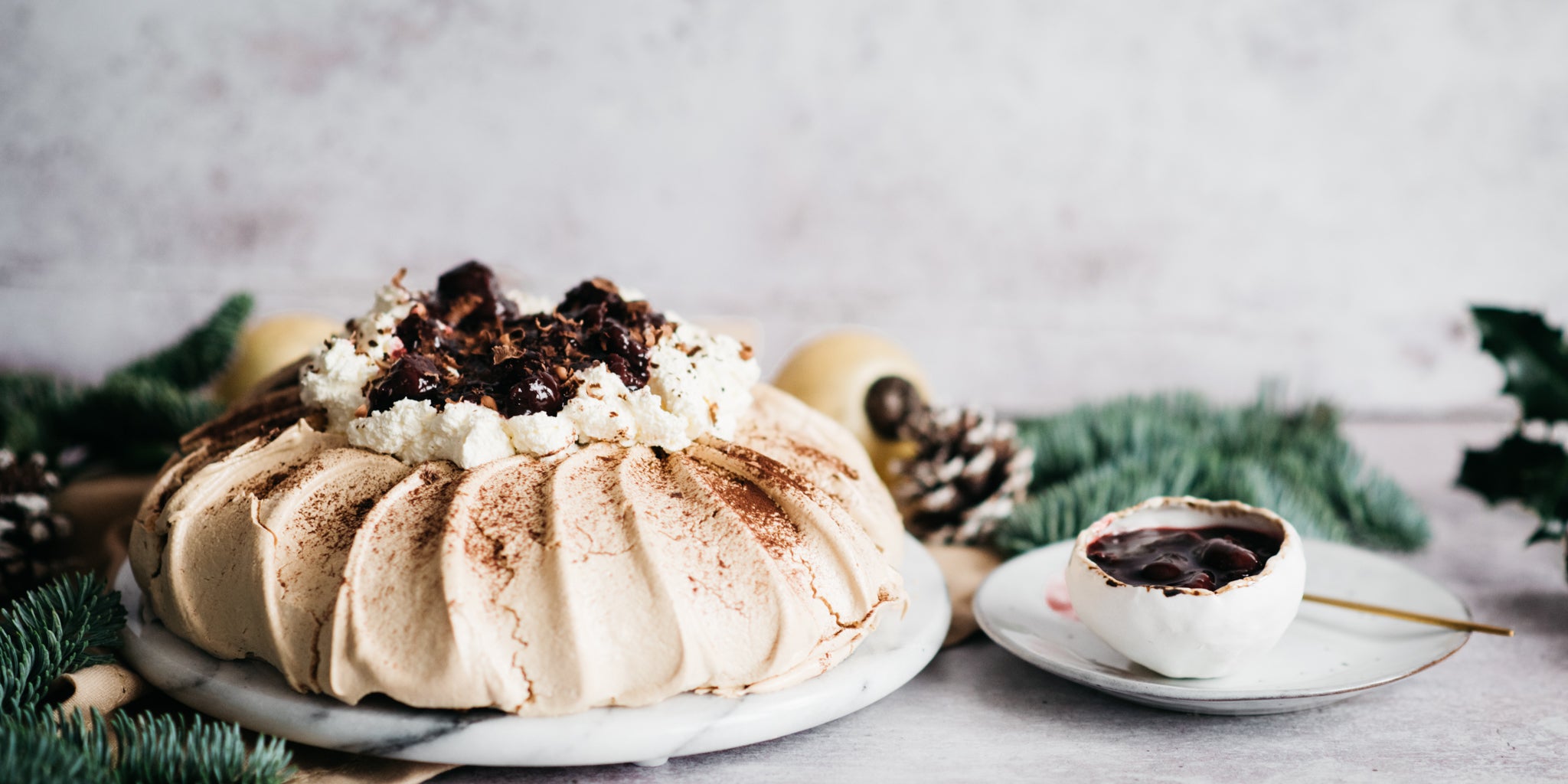 Black Forest Pavlova next to pine cones and leaves, with a bowl of cherry kirsch