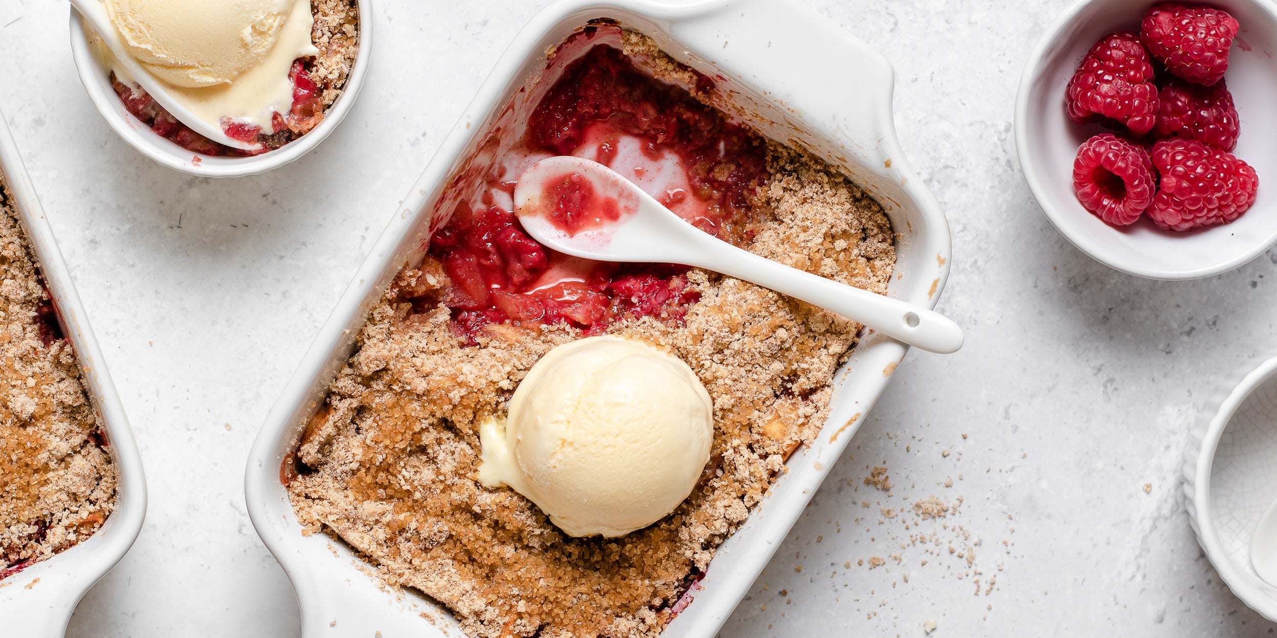 Top view of a golden topped Apple & Raspberry Crumble with a spoon dipping in for a portion, topped with a dollop of vanilla icecream