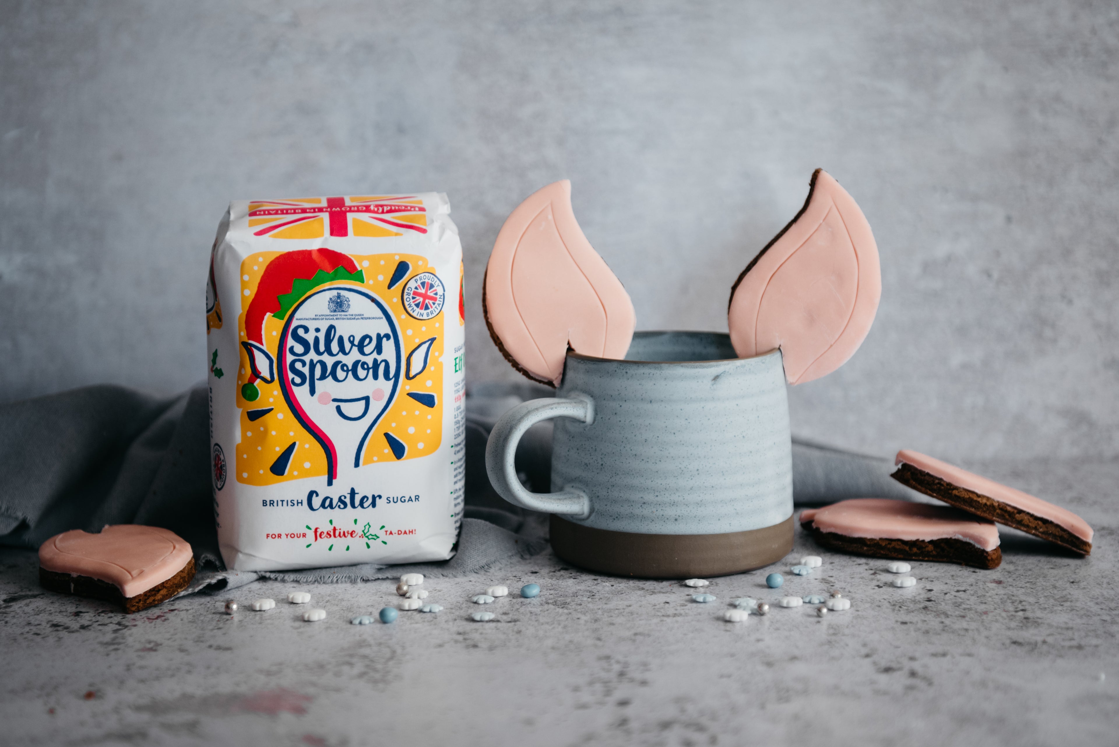 Elf Ears Gingerbread Cookies on the edge of a blue mug, next to a bag of Christmas edition packaging silver spoon caster sugar
