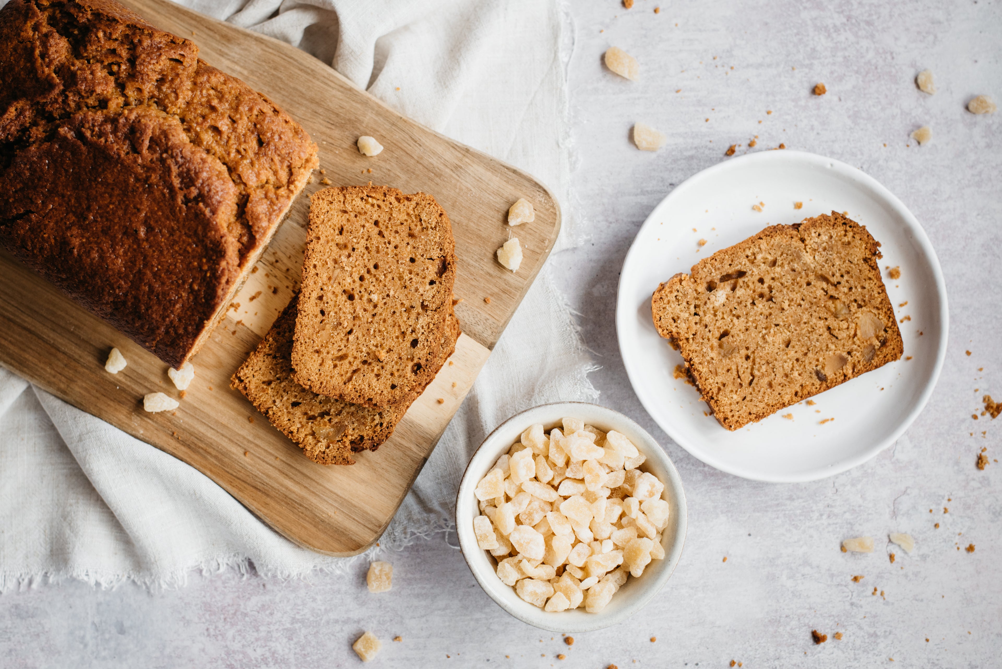 Top view of Gingerbread Loaf sliced on a wooden board, with a bowl of crystallised ginger, and a plate with a slice of Gingerbread Loaf