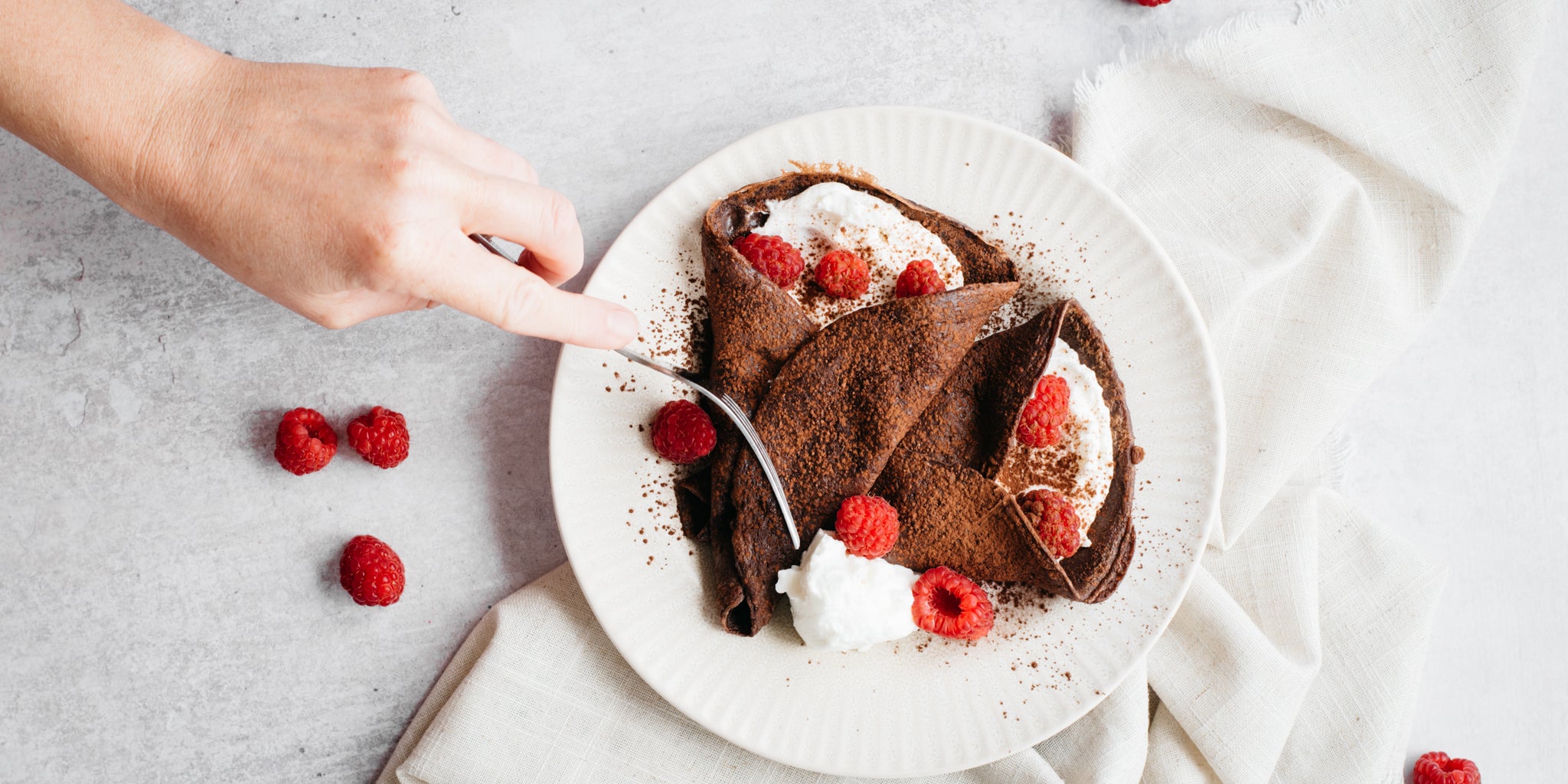 White plate with two chocolate crepes and raspberries. Hand reaching in with a fork