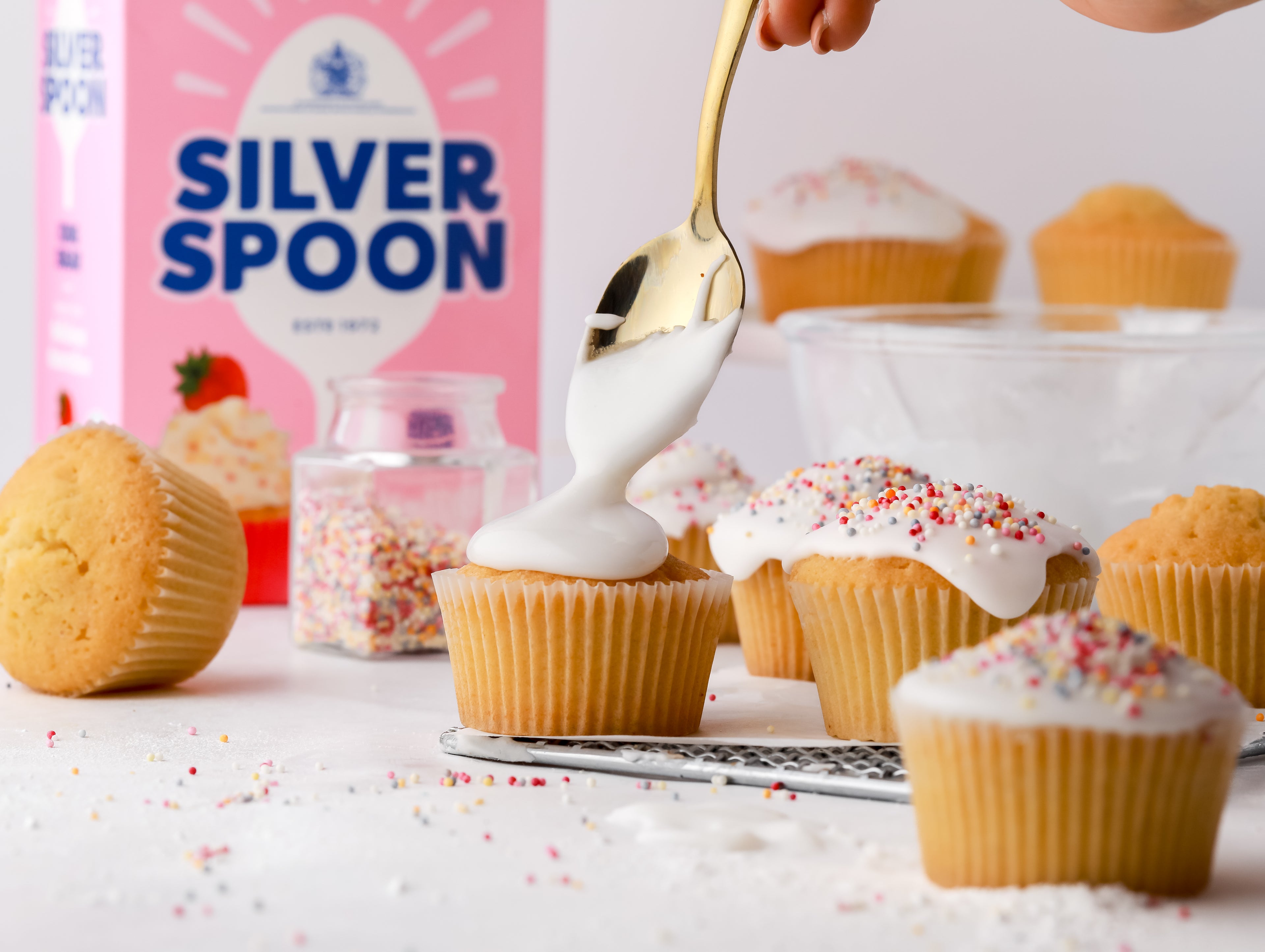 Icing being spooned on top of cupcakes with a jar of sprinkles and pack of icing sugar beside