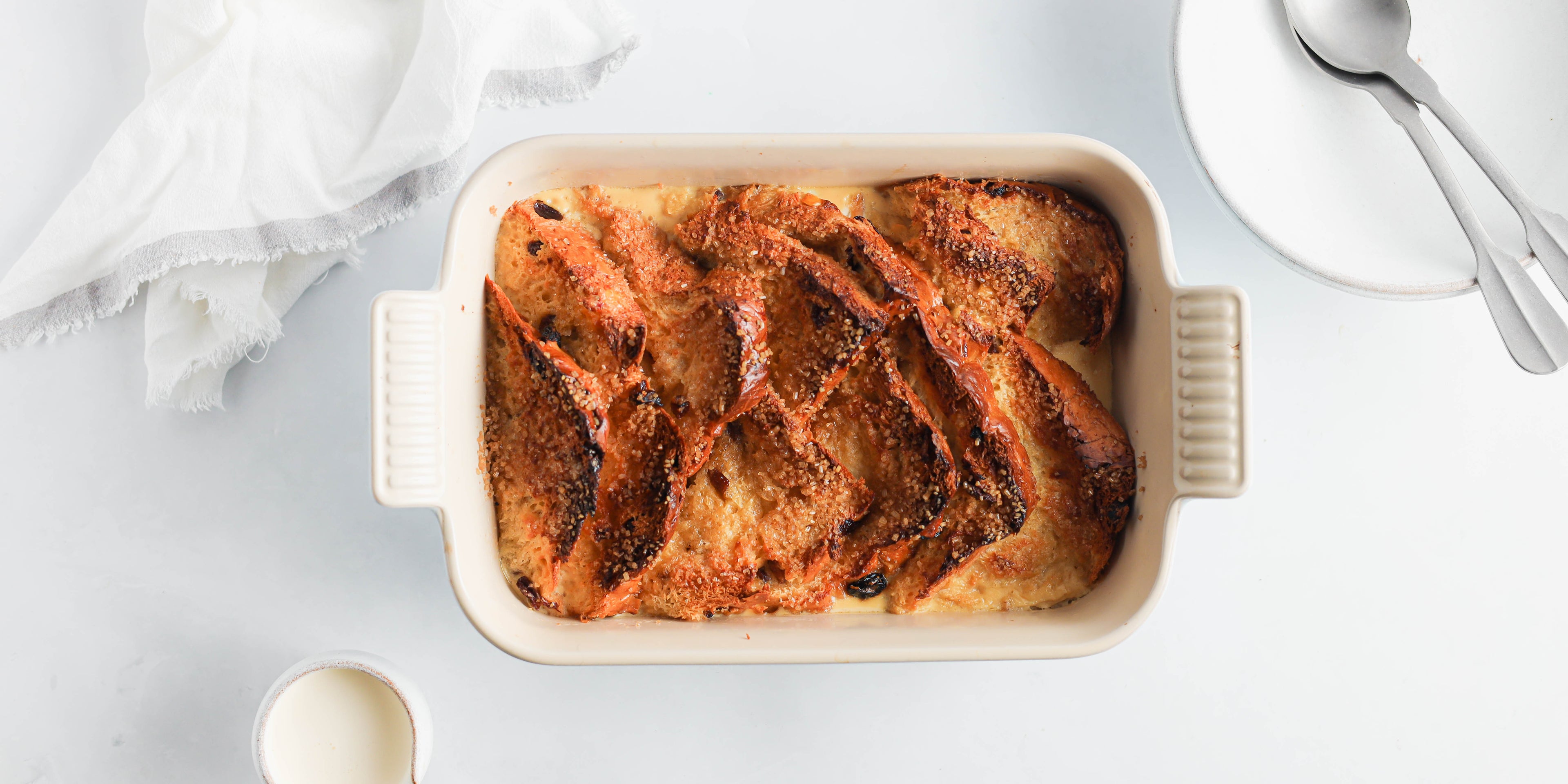 Top view of a golden topped Panettone Bread & Butter Pudding next to a jug of cream and spoons to serve