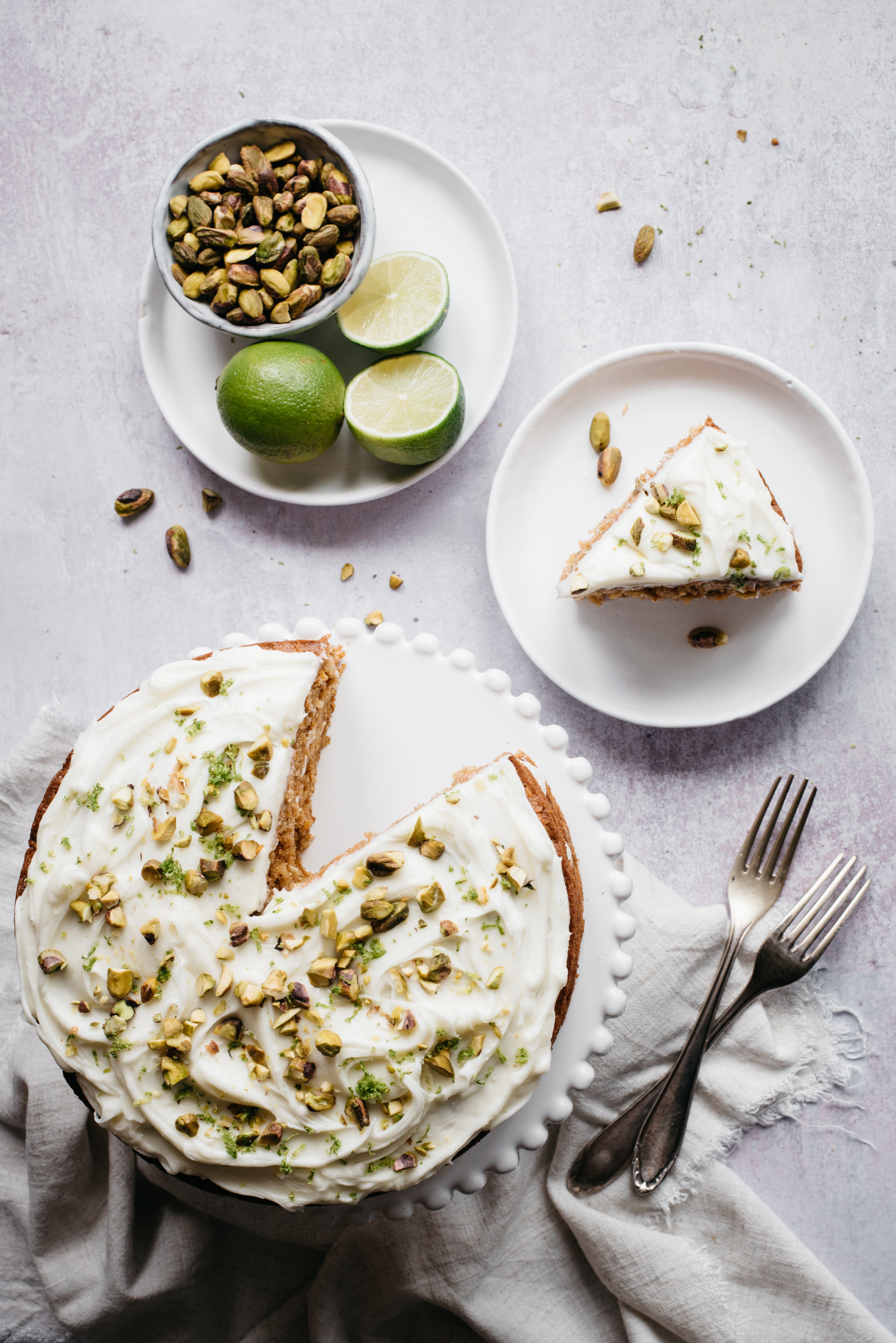 Over the top shot of cake with slice removed. 2 forks pistachios and limes