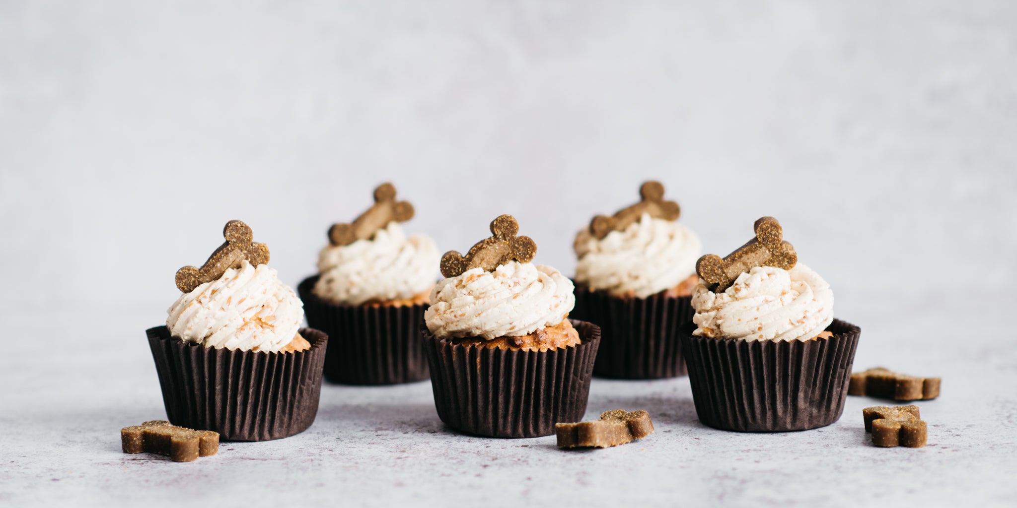 5 cupcakes in brown cases lined with frosting and a dog treat topping. Dog treats scatted around the base