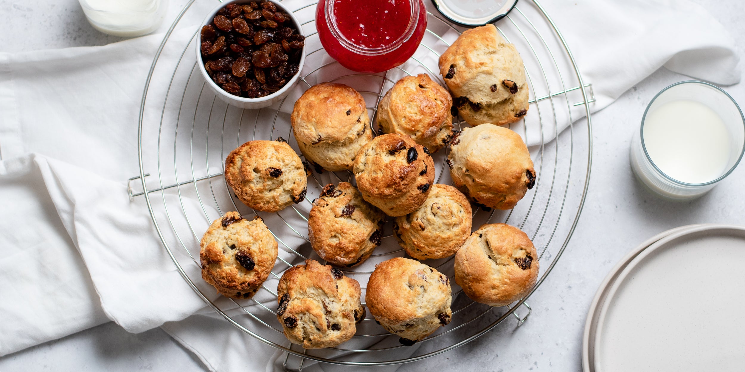 Top view of a fresh batch of Fruit Scones on a wire cooling rack, next to a bowl of jam, and sultanas