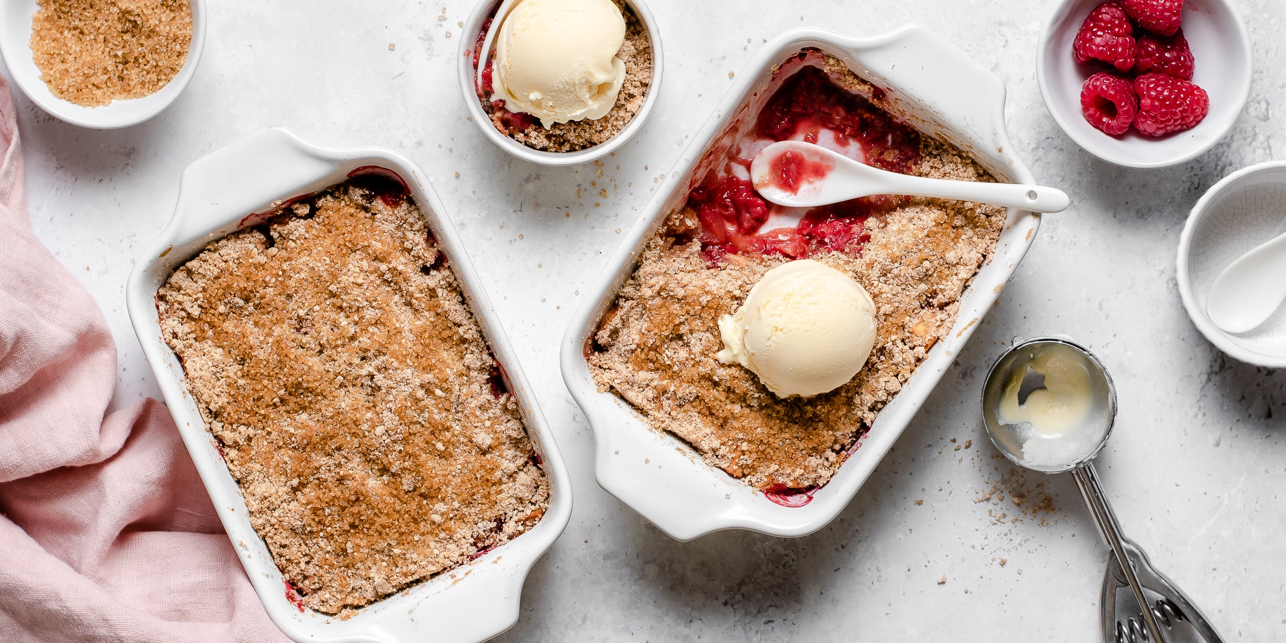 Top view of a golden topped Apple & Raspberry Crumble with a spoon dipping in for a portion, topped with a dollop of vanilla icecream
