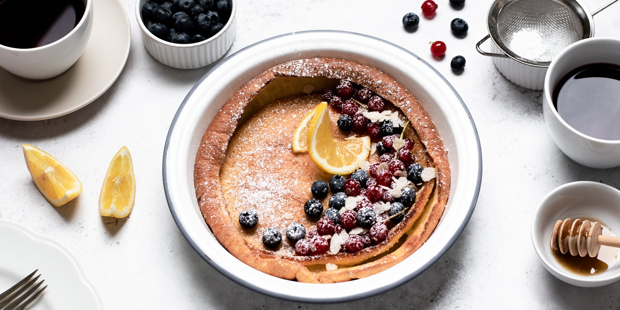 Dutch Baby Pancakes drizzled in honey, topped with berries and lemon, next to cups of coffee, a honey drizzler and ramekin of blueberries