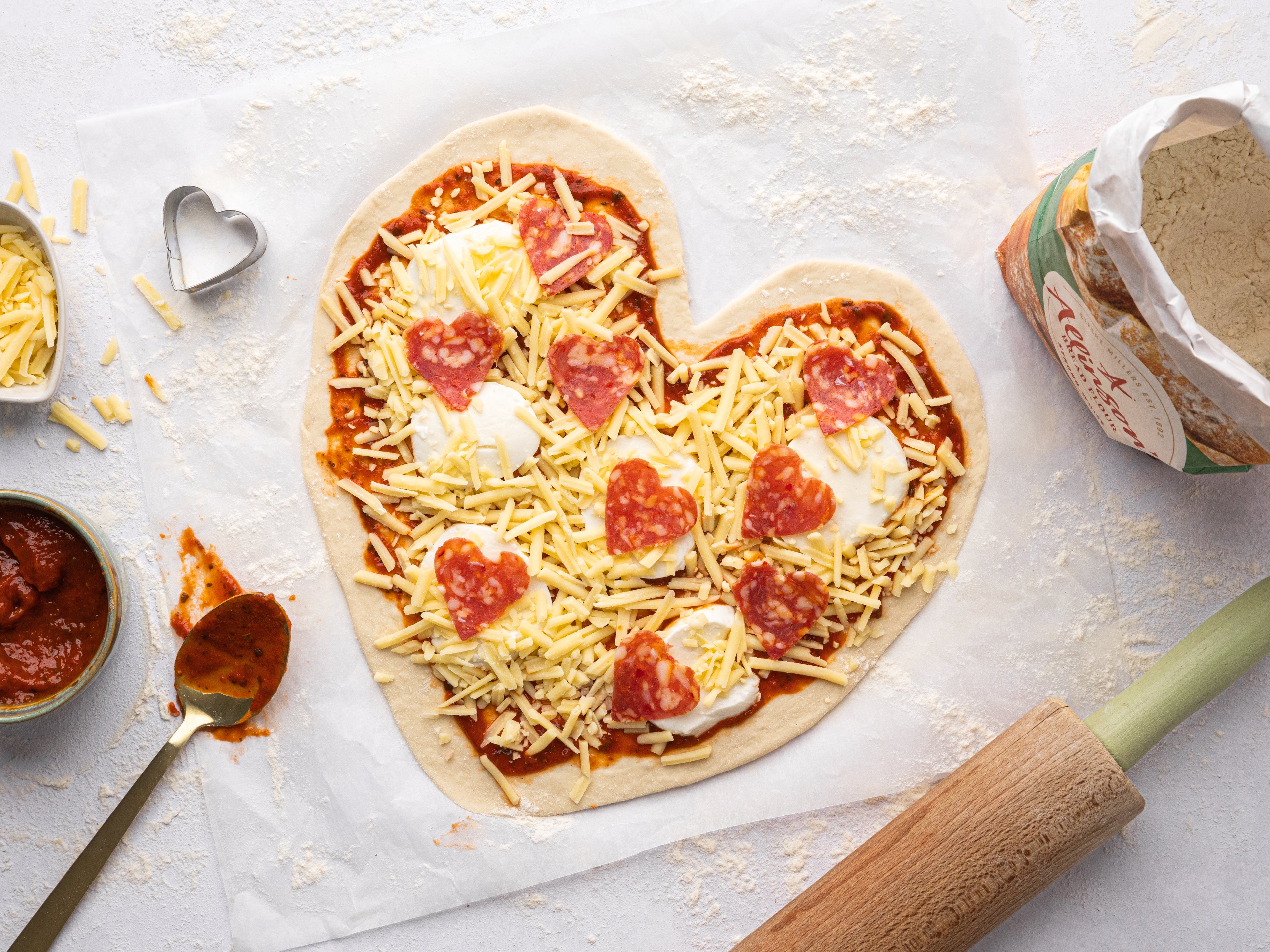 Heart shaped pizza topped with heart shaped pepperoni beside a bag of flour and messy kitchen scene
