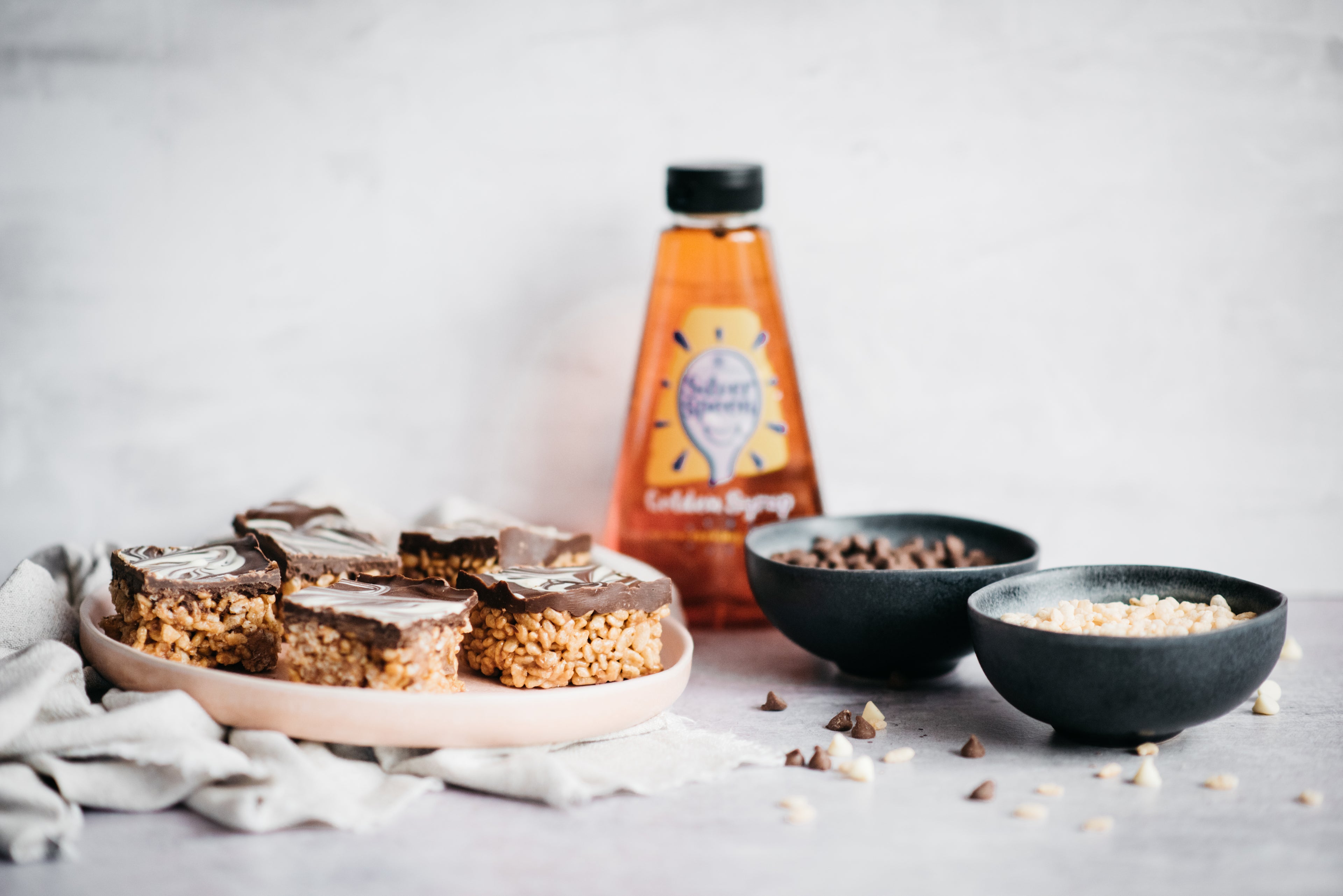 Chocolate Mars Bar Cakes on a plate next to bowls of cereal and chocolate chips, and a bottle of golden syrup