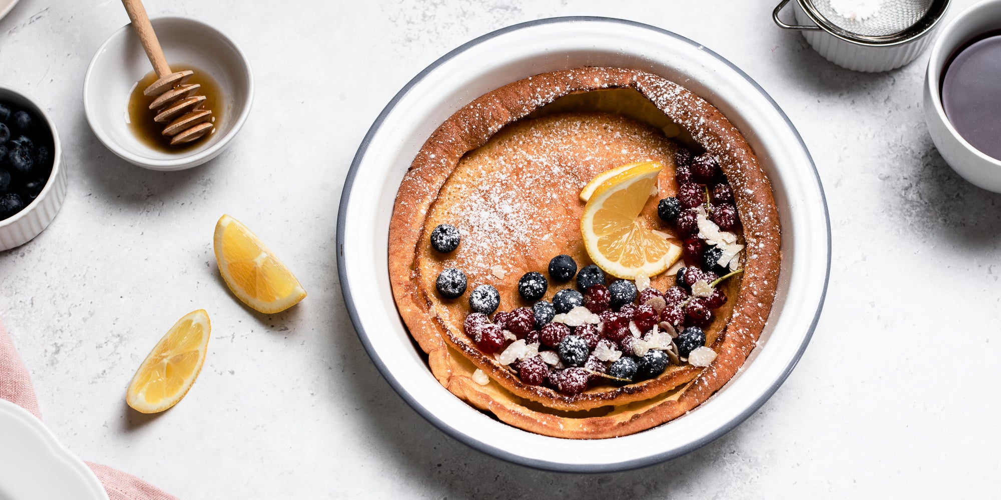 Top view of Dutch Baby Pancakes drizzled in honey, topped with berries and lemon, next to cups of coffee, a honey drizzler and ramekin of blueberries