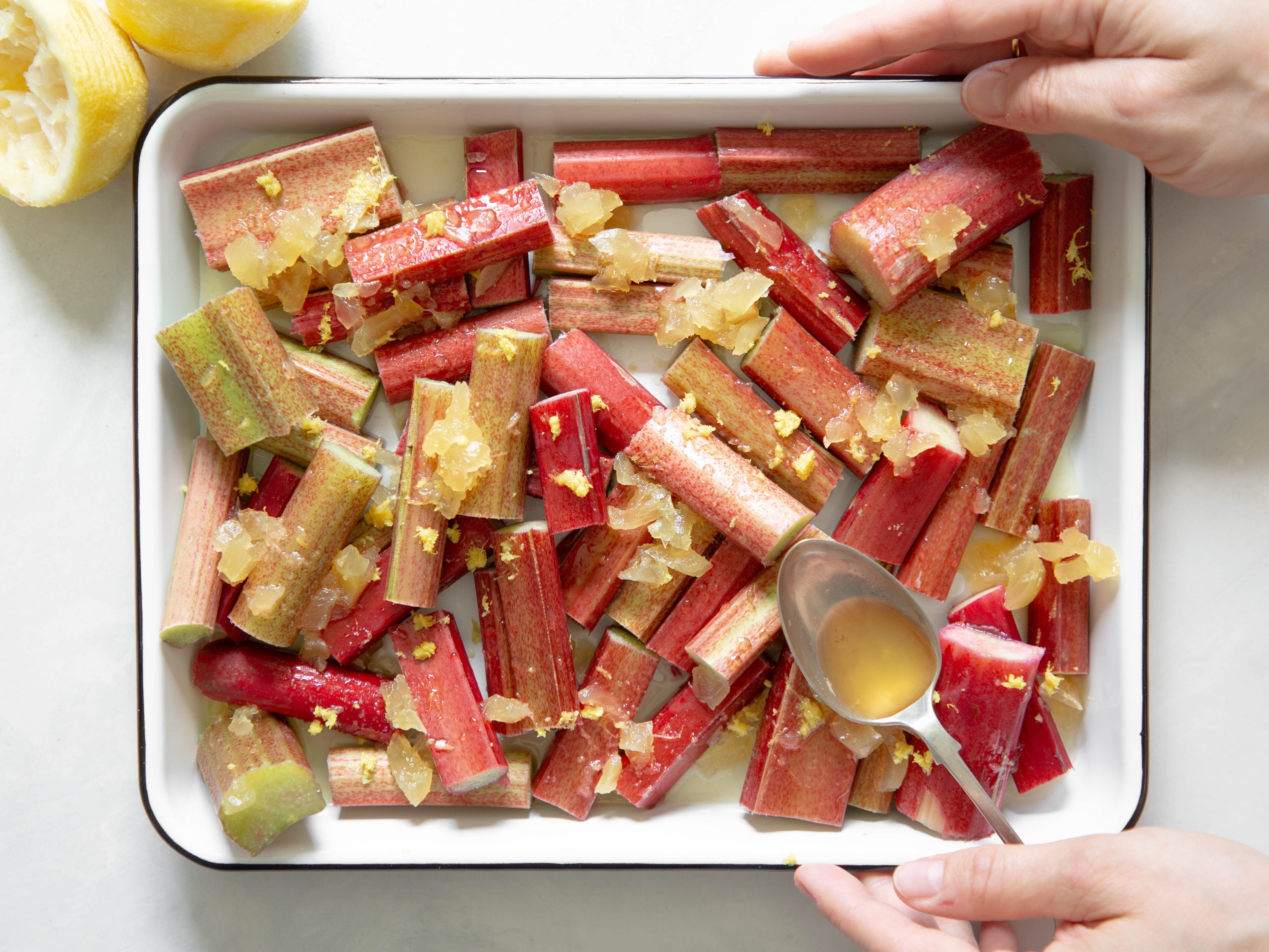 Baking dish full of chopped fresh rhubarb and two hands holding the right hand side. In the top left corner there is a squeezed lemon