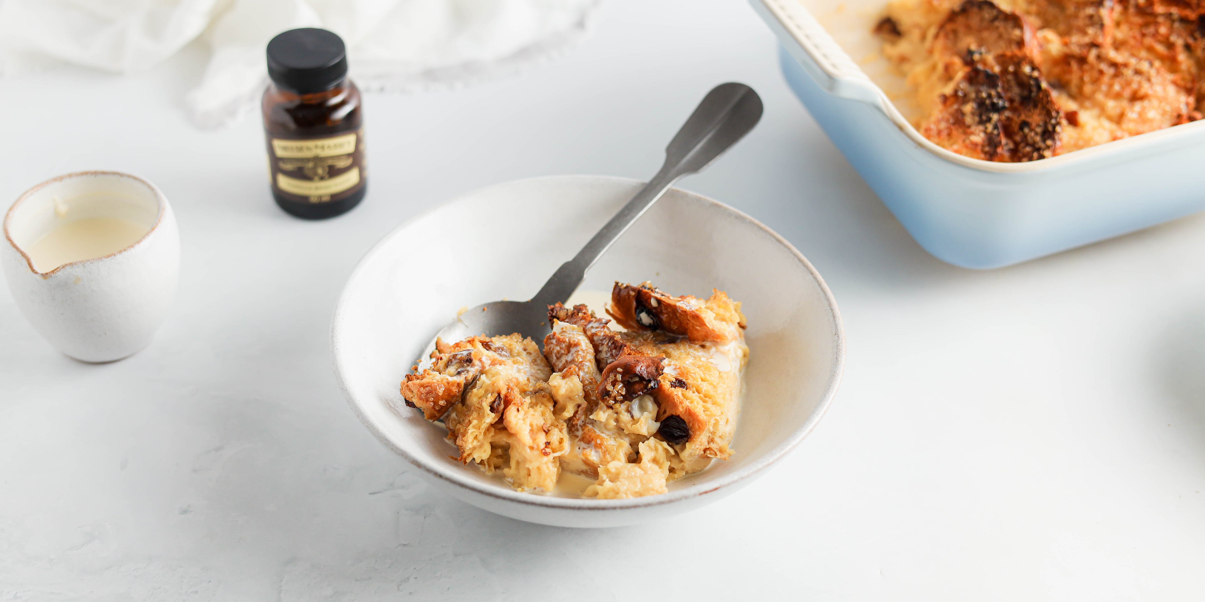 A bowl of Panettone Bread & Butter Pudding with a spoon ready to serve. Bottle of Nielsen-Massey vanilla extract in the background