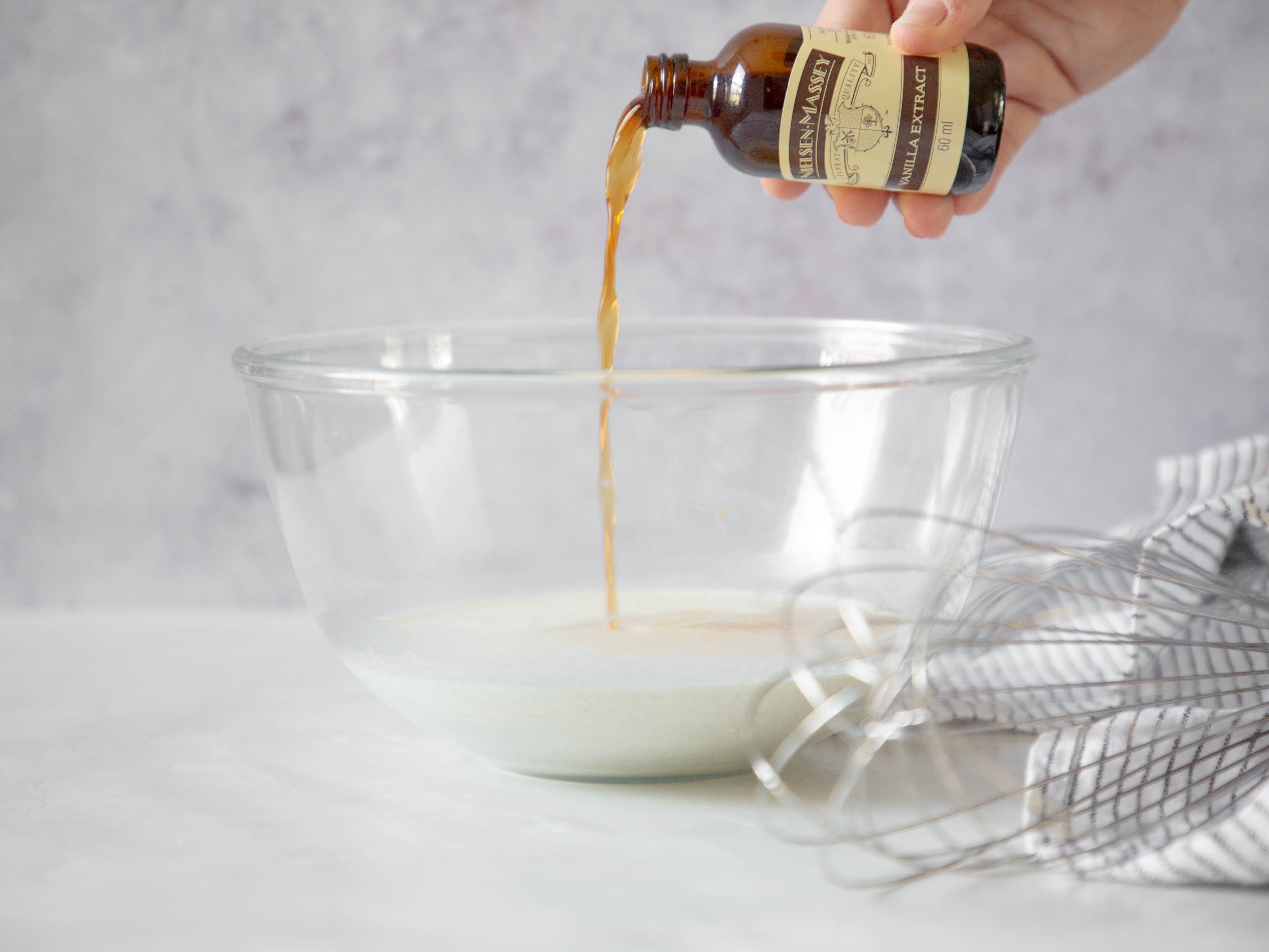 Hand pouring a bottle of vanilla extract into a glass bowl with a whisk and tea towel in the forefront
