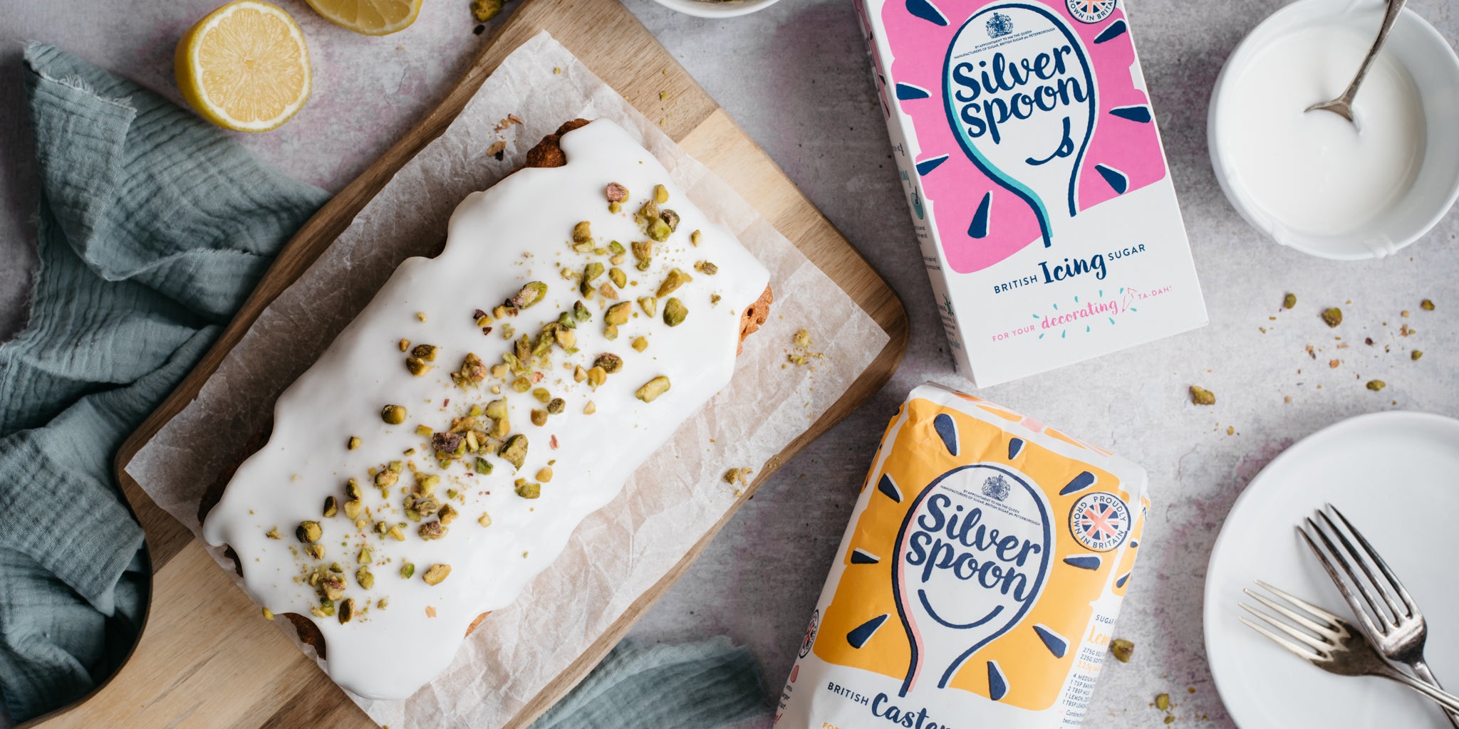 Top view of Gluten Free Vegan Lemon Drizzle Cake topped with nuts, next to a box of Silver Spoon Caster and Icing Sugar