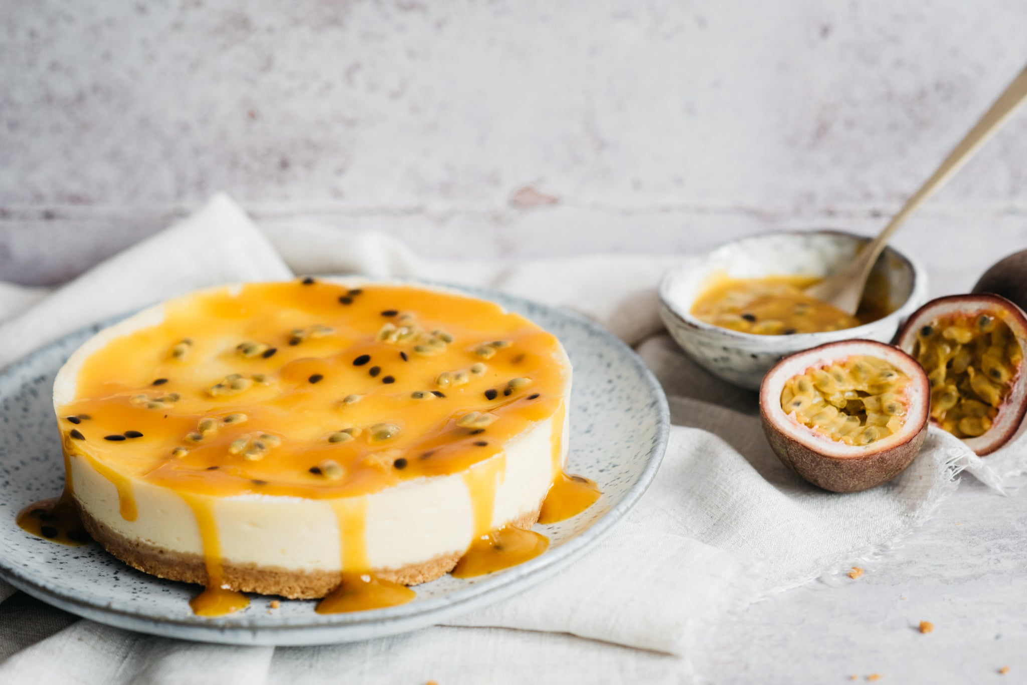 Cheesecake on a plate with passion fruit topping drizzled. Bowl of passionfruit sauce and spoon in background with passion fruits
