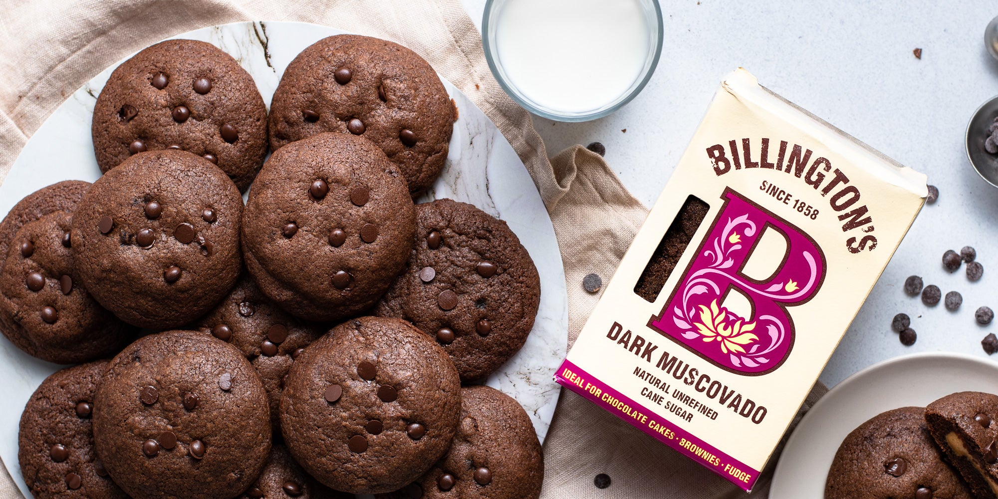 A batch of Chocolate & Peanut Butter Stuffed Cookies on a plate, next to a Billington's Dark Muscovado Sugar and a glass of milk