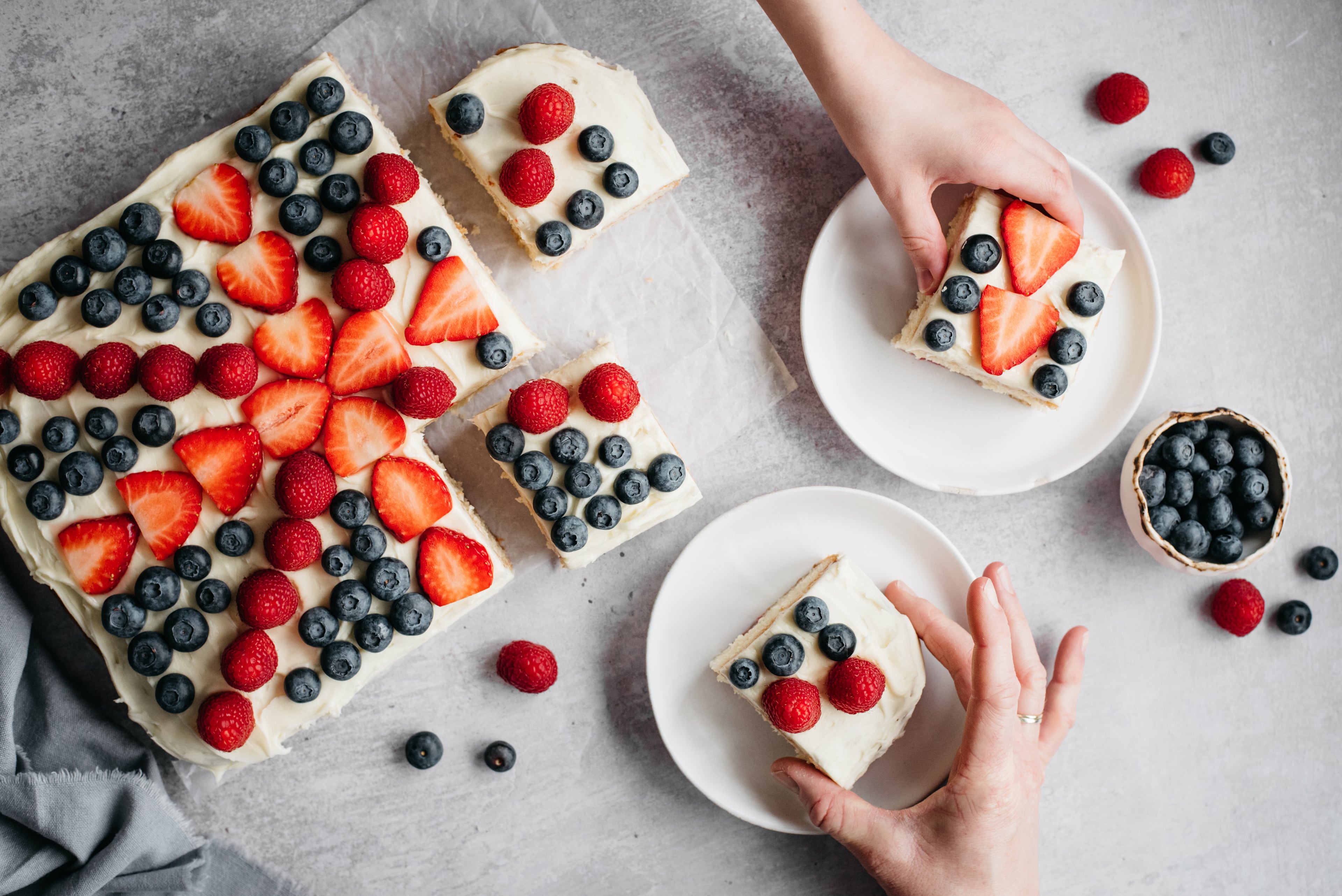Top view of Union Jack Traybake with sliced served on plates, and hands reaching for a piece of cake topped with fresh berries for the Jubilee