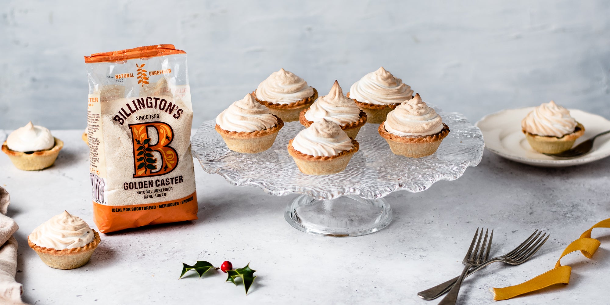 Meringue Topped Mince Pies on a glass cake stand next to a bag of Billington's Golden caster sugar