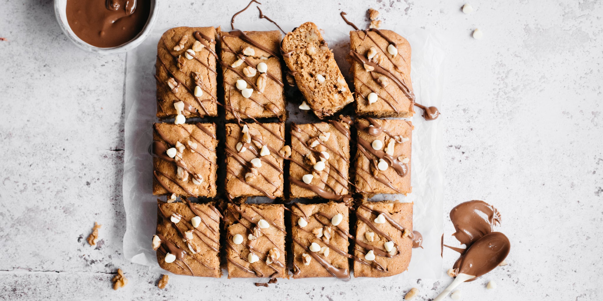 Overhead shot of peanut butter blondies cut into 12 chunks, one chunk turned on its side. A bowl of melted chocolate beside and a spoon