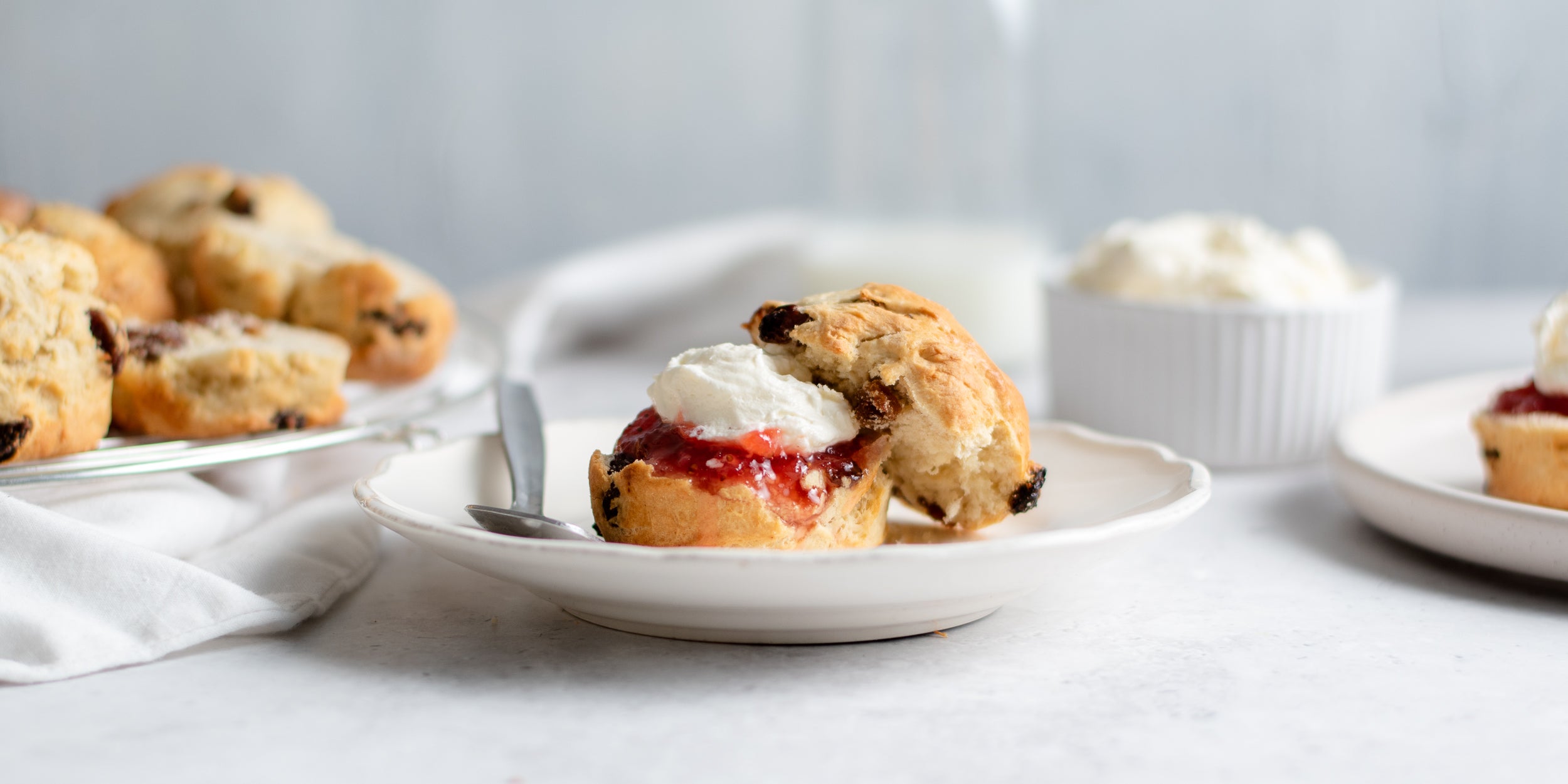 Close up of Fruit Scones served on a plate, cut in half with a dollop of cream and jam. With a ramekin of whipped cream in the background