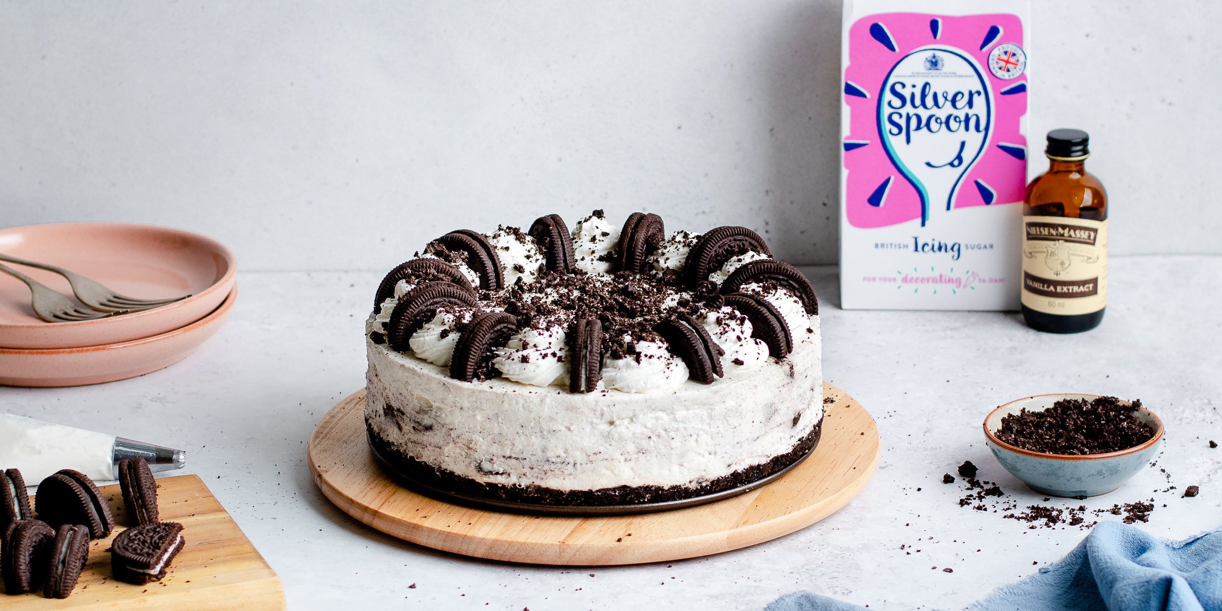 Oreo cheesecake on a wooden circular board. Vanilla extract and sugar pack in background. Plates and ingredients beside