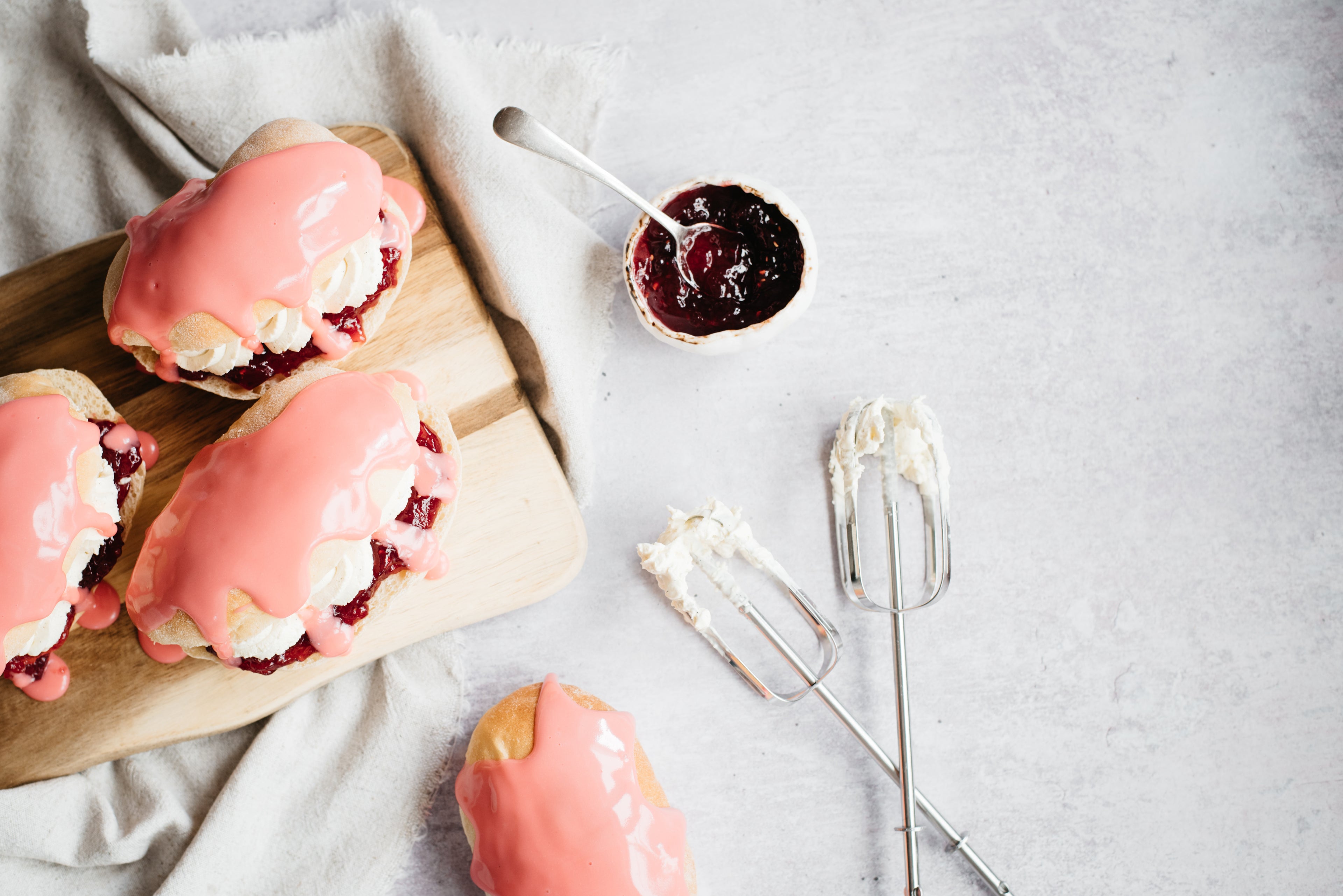 Top view of Iced Finger Buns on a serving board, next to a bowl of jam and whisks covered in Iced Finger Bun mixture