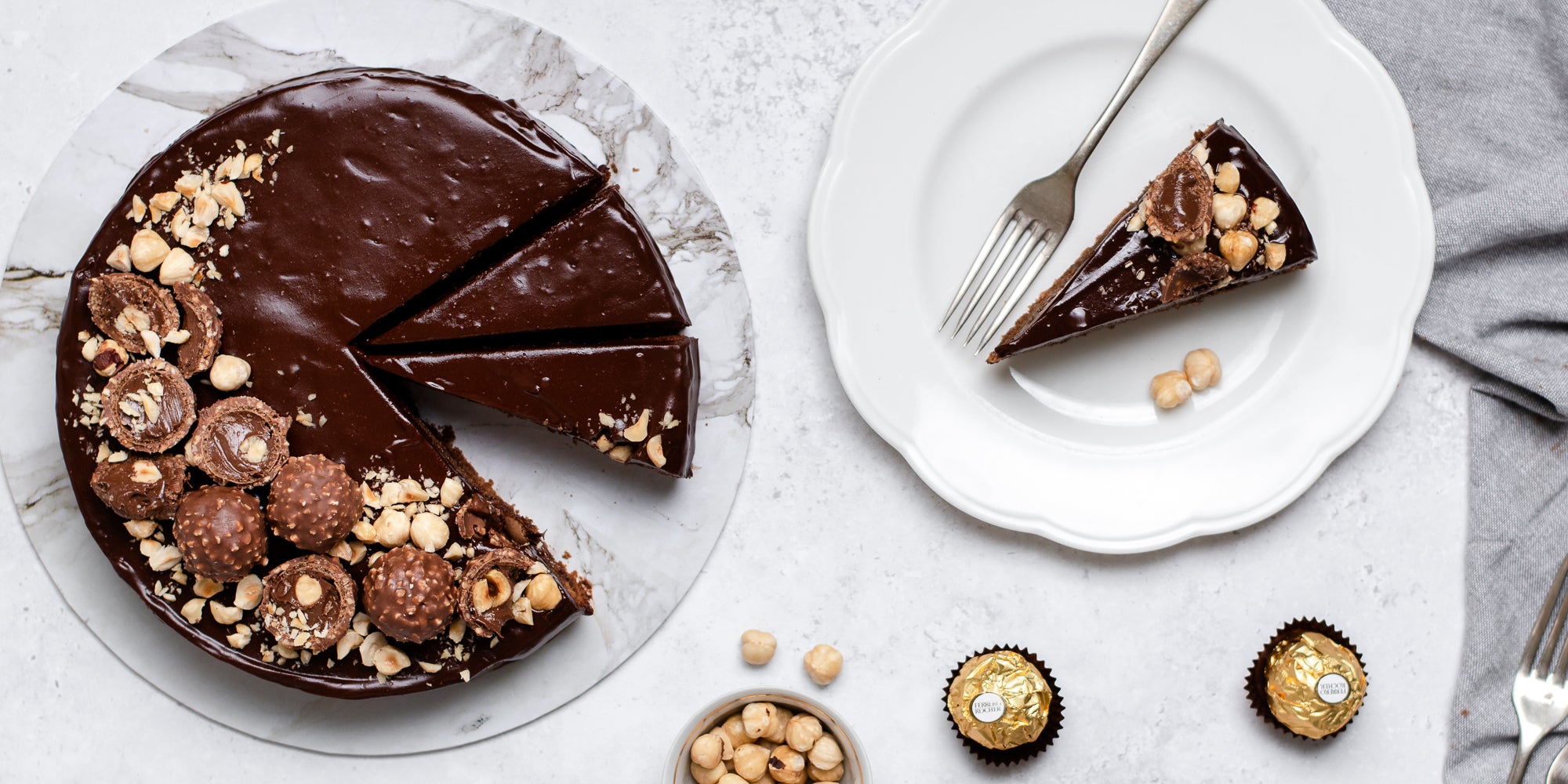 Top view of Gluten Free Chocolate Truffle Sachertorte sliced into servings, with a slice served on a plate next to a fork. Smothered in chocolate icing, Sprinkled with Ferrero Rochers and chopped hazelnuts.