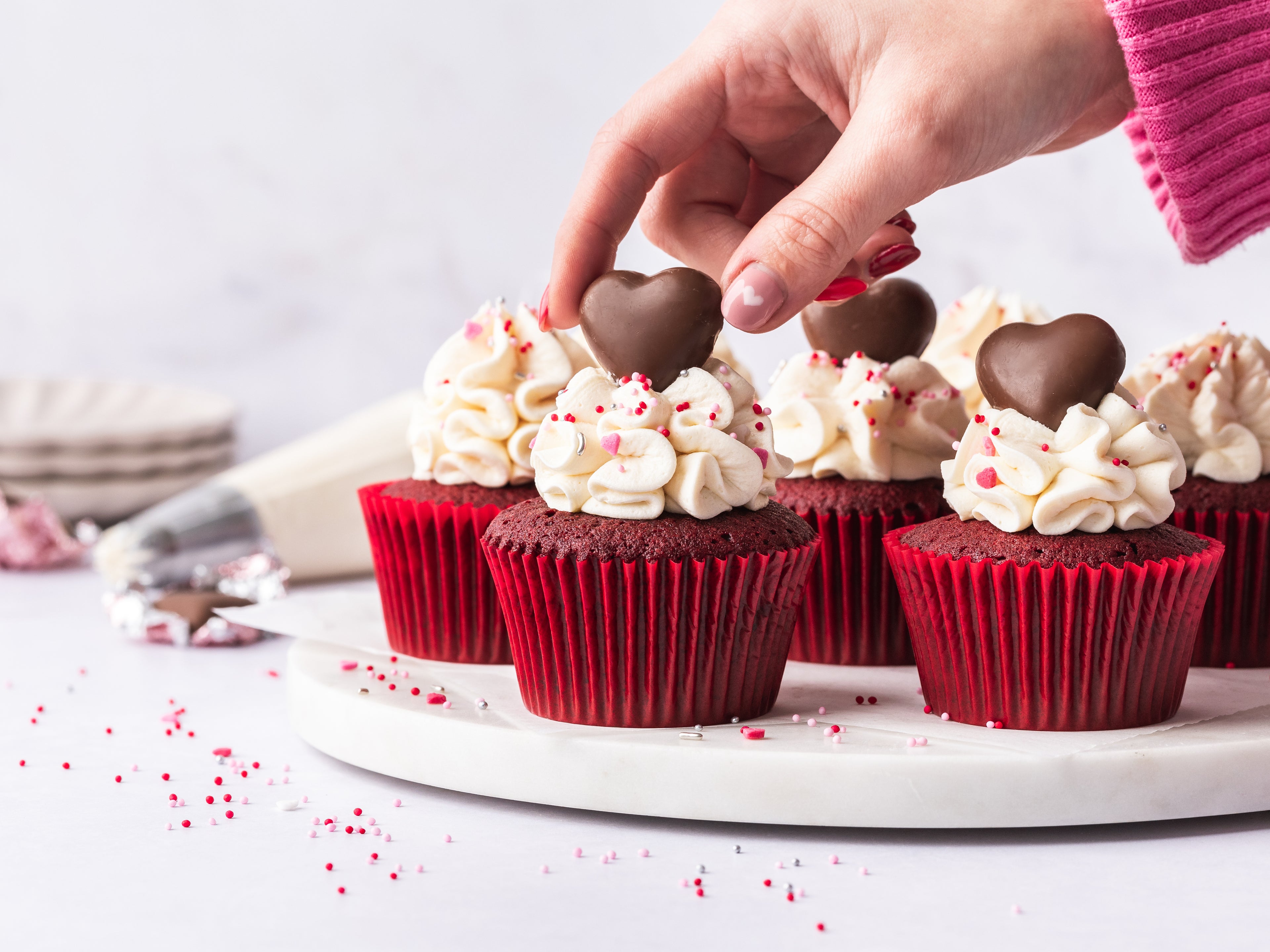 Hand placing a heart shaped decoration on the top of the red velvet cupcakes