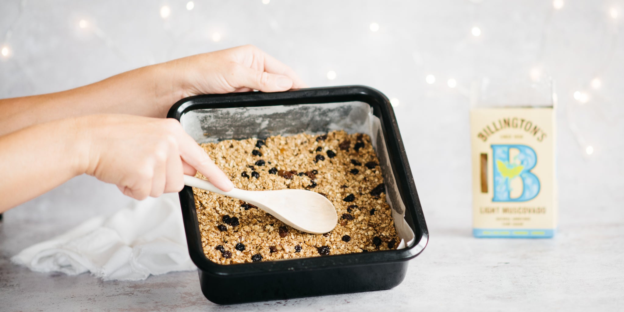 Hand pushing down flapjack mixture with a wooden spoon and sugar pack in background